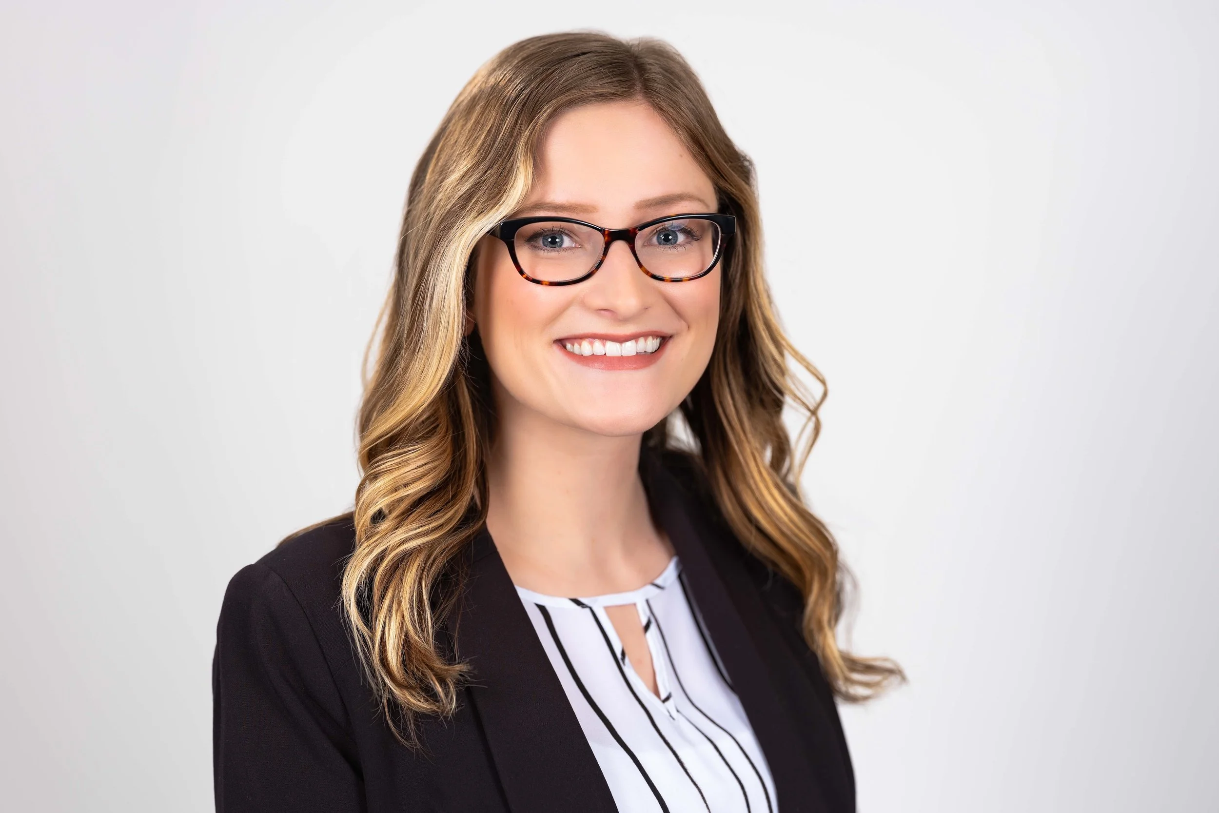 Professional Corporate Headshot by Minneapolis Photographer Jay Cupcake. Portrait of a smiling young woman with wavy brown hair, glasses, wearing a black blazer and a white and black striped blouse, against a plain white background.