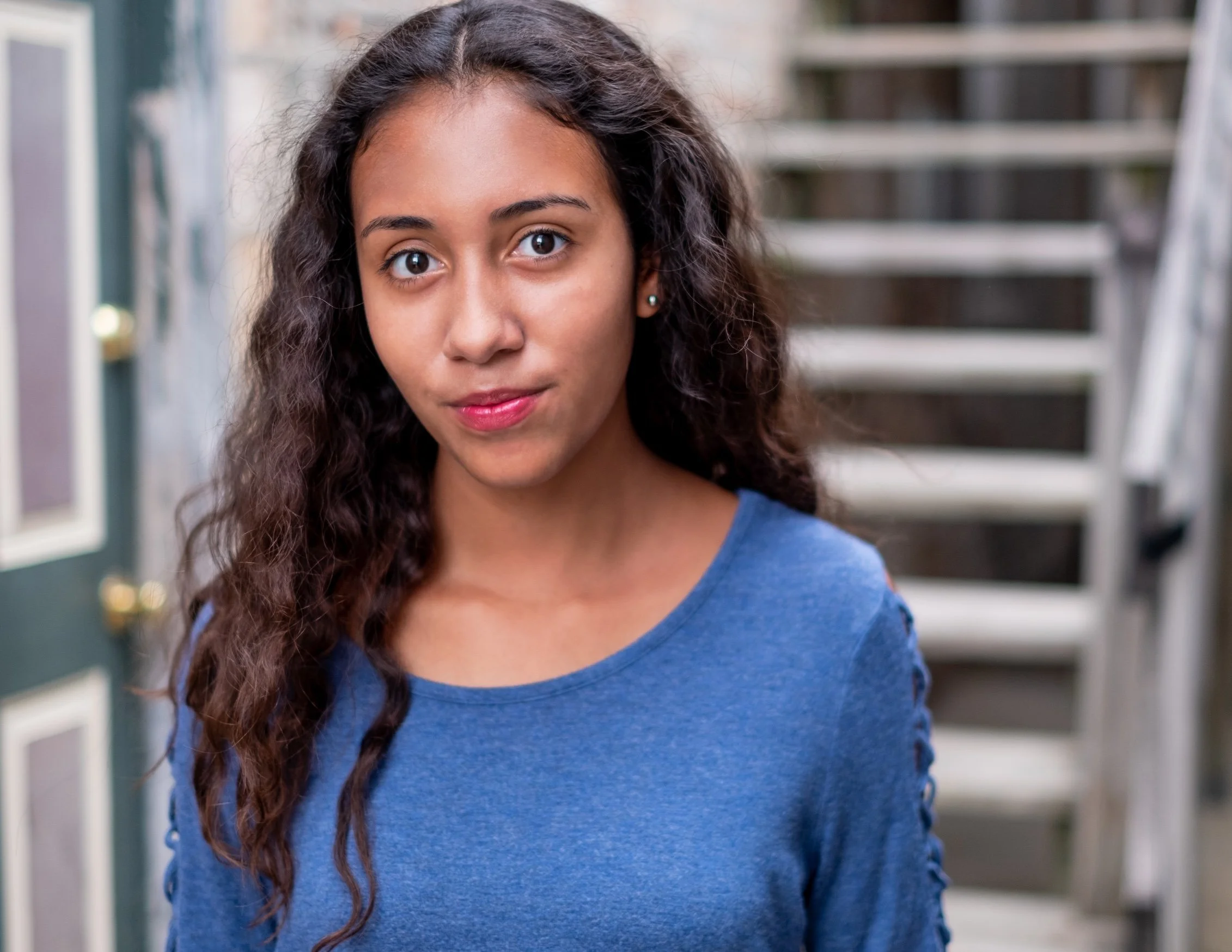 Professional Actor Headshot by Saint Paul Photographer Jay Cupcake. A young woman with dark, curly hair and brown eyes looking at the camera, standing outdoors near stairs and a door.
