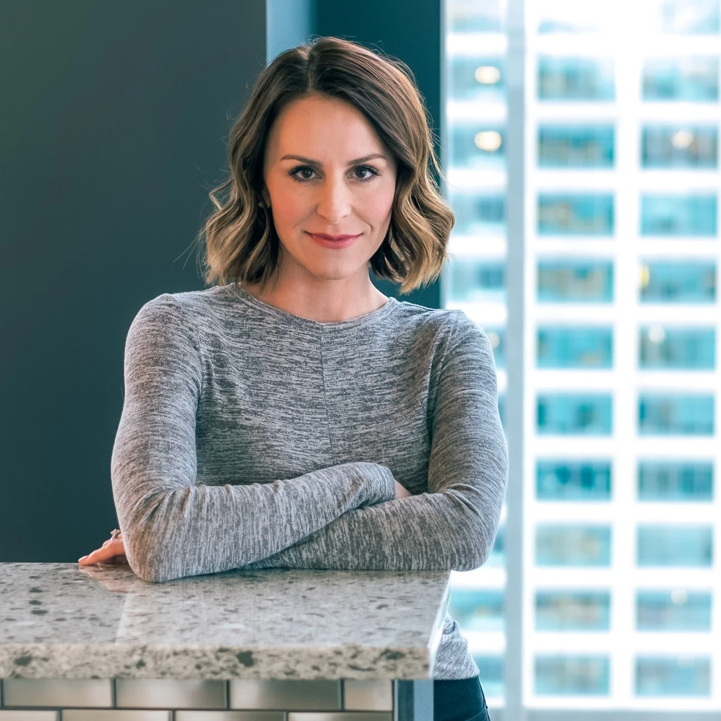 Professional Portrait Photography by Minneapolis Photographer Jay Cupcake. A woman with shoulder-length wavy brown hair, wearing a gray long-sleeve shirt, crossing her arms and smiling while standing in a modern office with large windows and cityscap