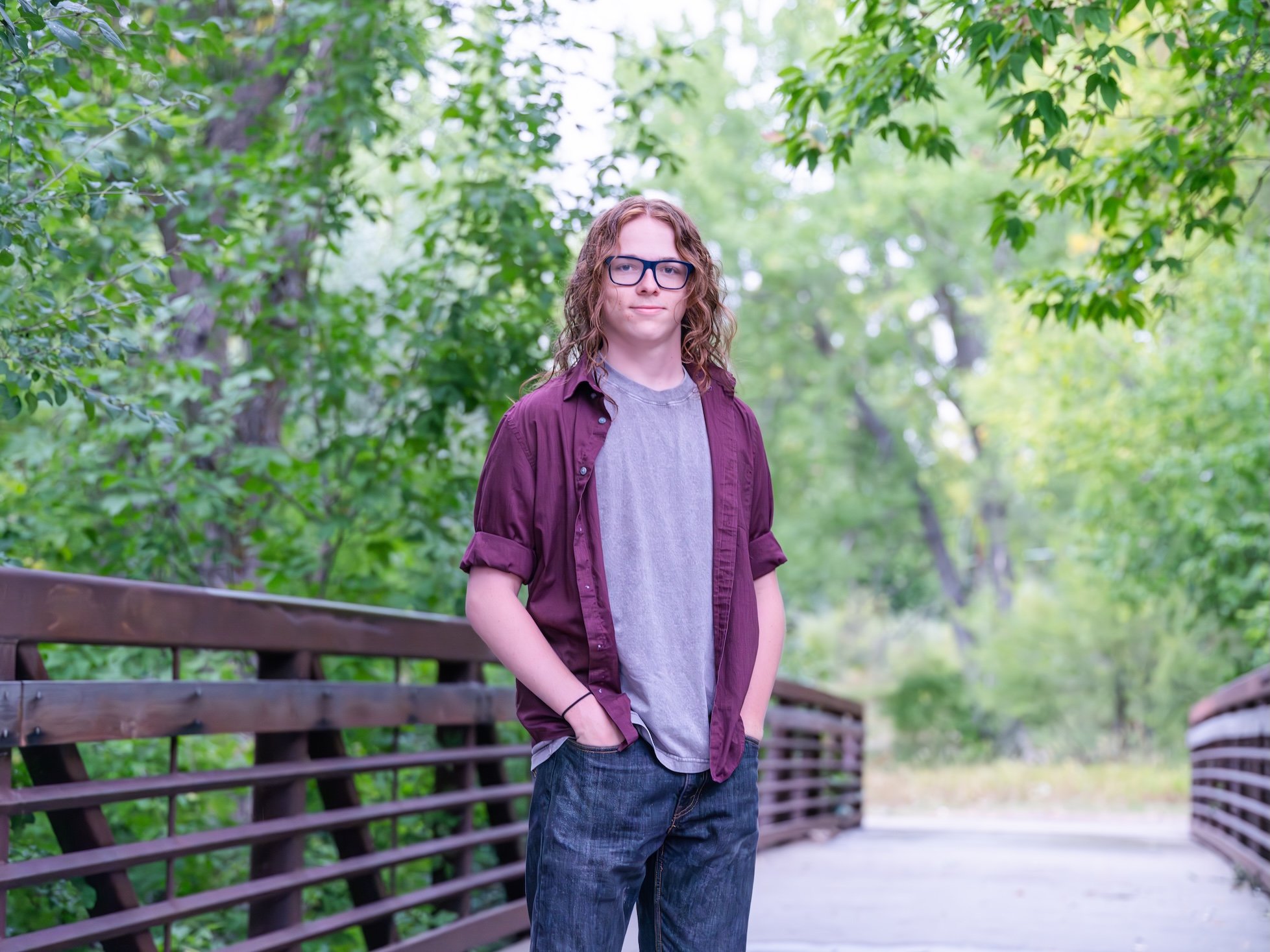 Professional Portrait Photography by Minneapolis Photographer Jay Cupcake. A young person with shoulder-length curly hair, glasses, wearing a gray t-shirt and purple button-up shirt, standing on a wooden bridge surrounded by greenery.