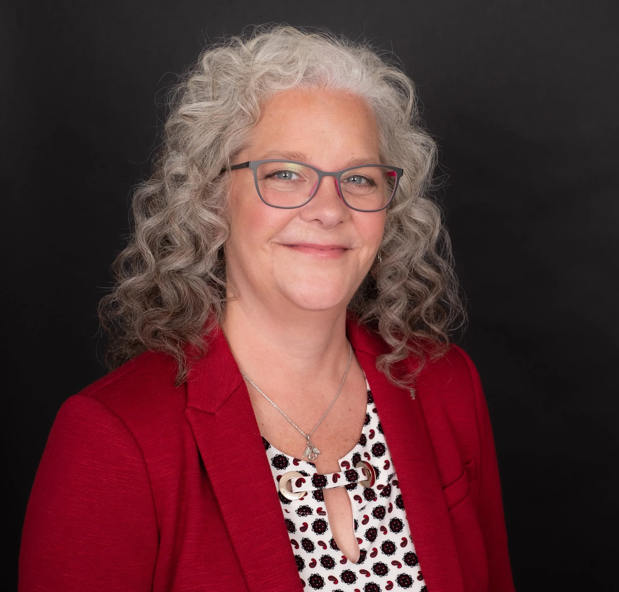 Professional Corporate Headshot by Saint Paul Photographer Jay Cupcake. A middle-aged woman with curly gray hair, glasses, and a warm smile, wearing a red blazer over a patterned blouse, against a black background.