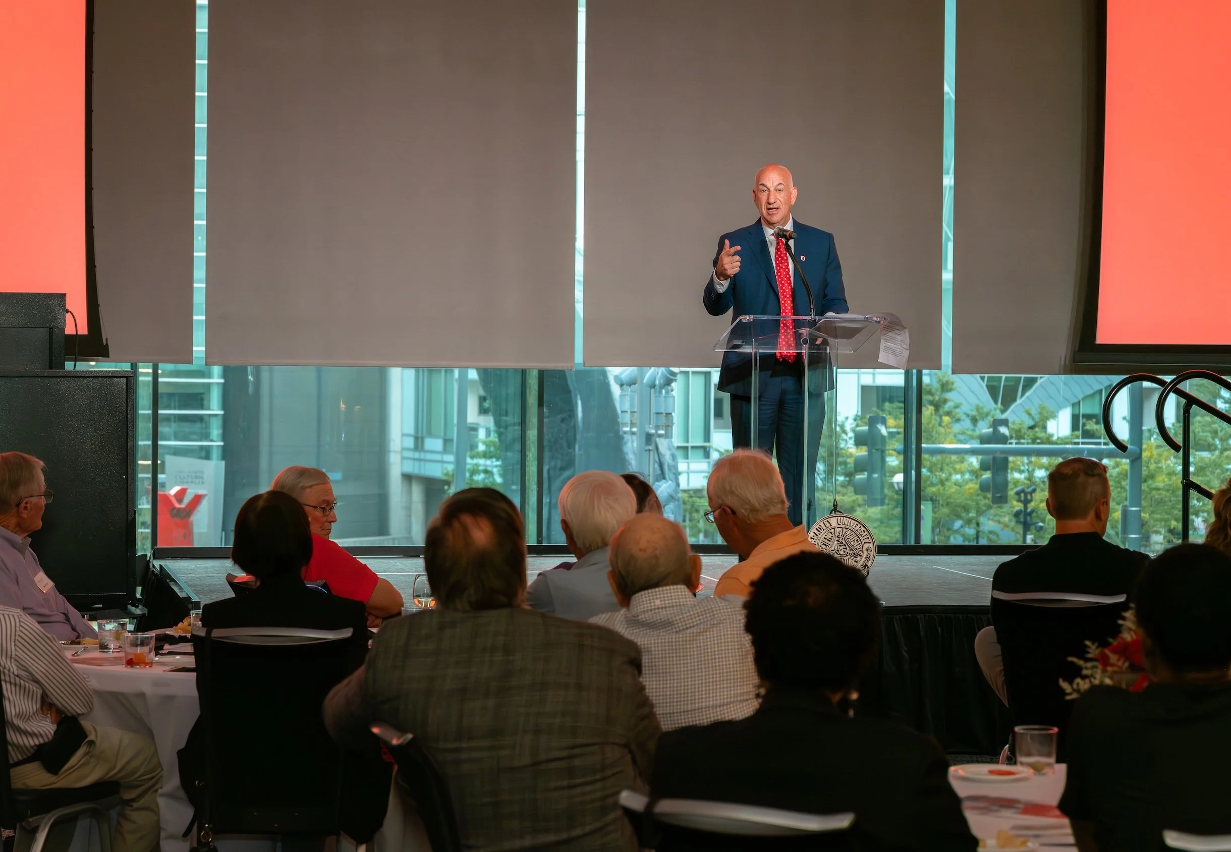 Professional Event Photography by Twin Cities Photographer Jay Cupcake. A man in a blue suit giving a speech at a podium during a conference or event with an audience seated at round tables. The background features large windows with cityscape views.
