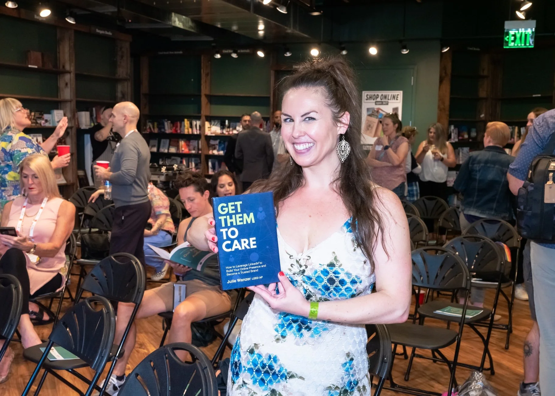 Professional Event Photography by Saint Paul Photographer Jay Cupcake. A woman in a white and blue dress holding a book titled "Get Them To Care" at a crowded indoor event, smiling and gesturing with her right hand. Other people are seated and standi