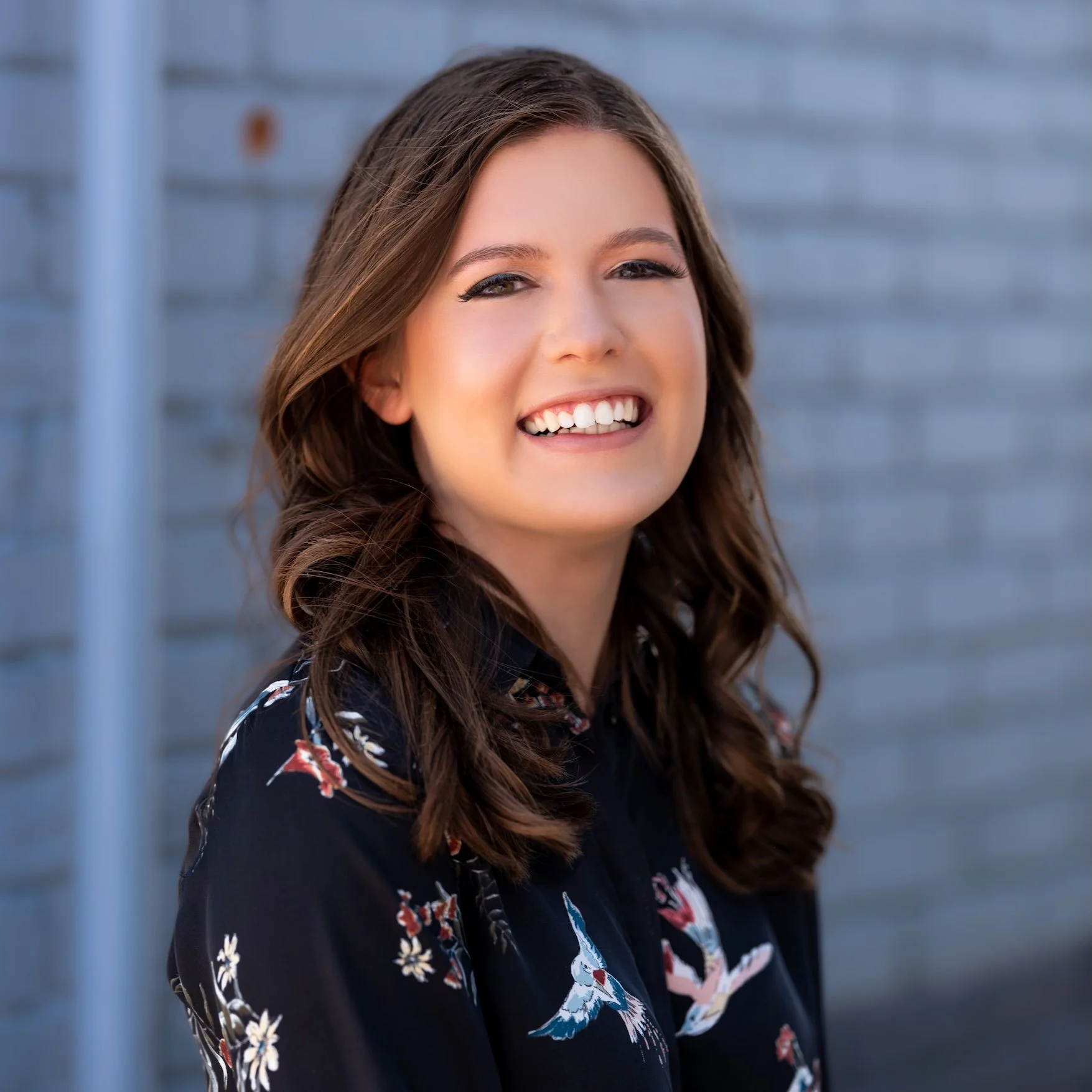 Professional Portrait Photography by Minneapolis Photographer Jay Cupcake. A young woman with brown, wavy hair smiling outdoors in front of a gray brick wall, wearing a dark blouse with floral and bird patterns.