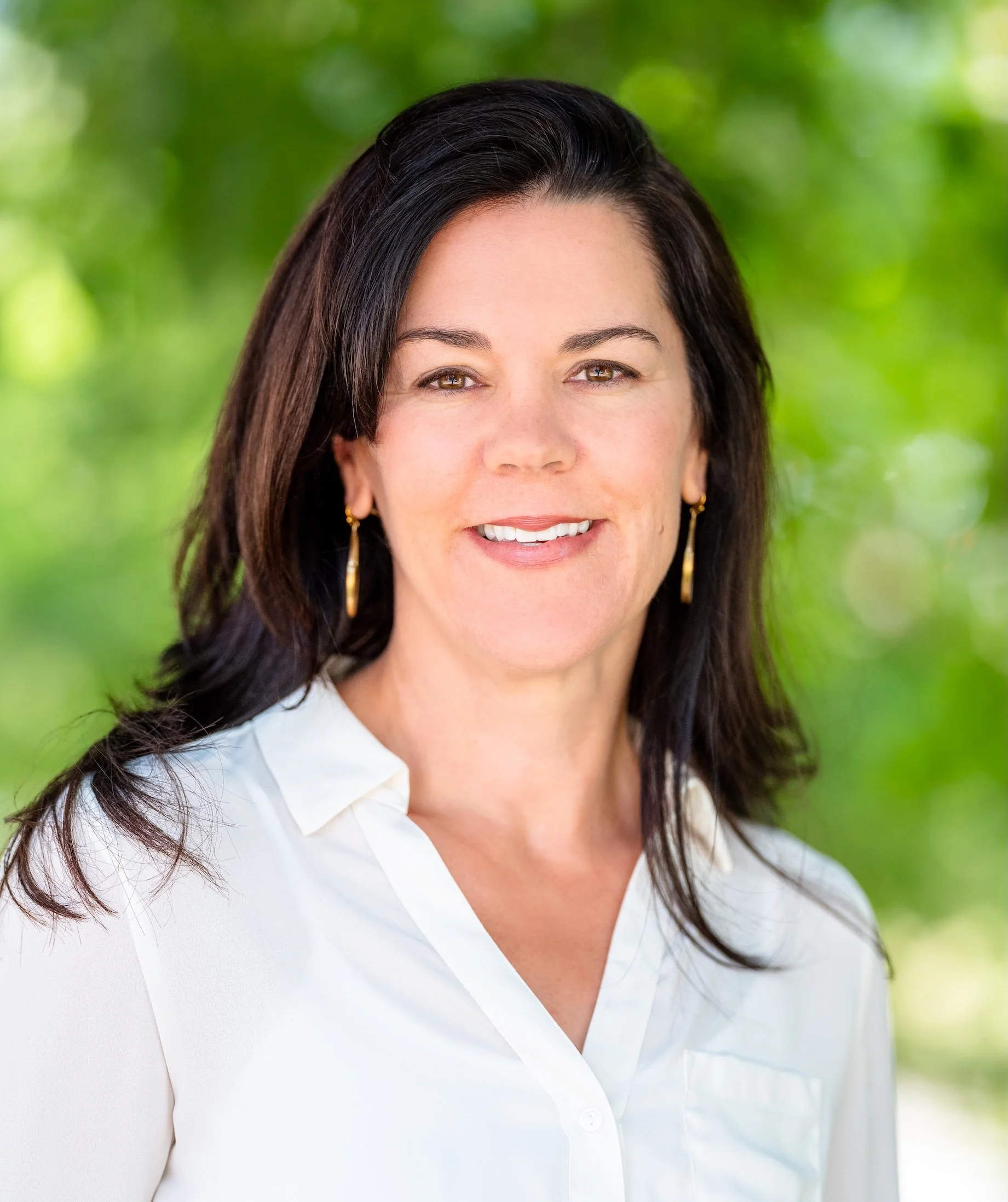 Professional Corporate Headshot by Minneapolis Photographer Jay Cupcake. A woman with long dark hair wearing a white shirt and gold earrings, smiling outdoors with green trees in the background.
