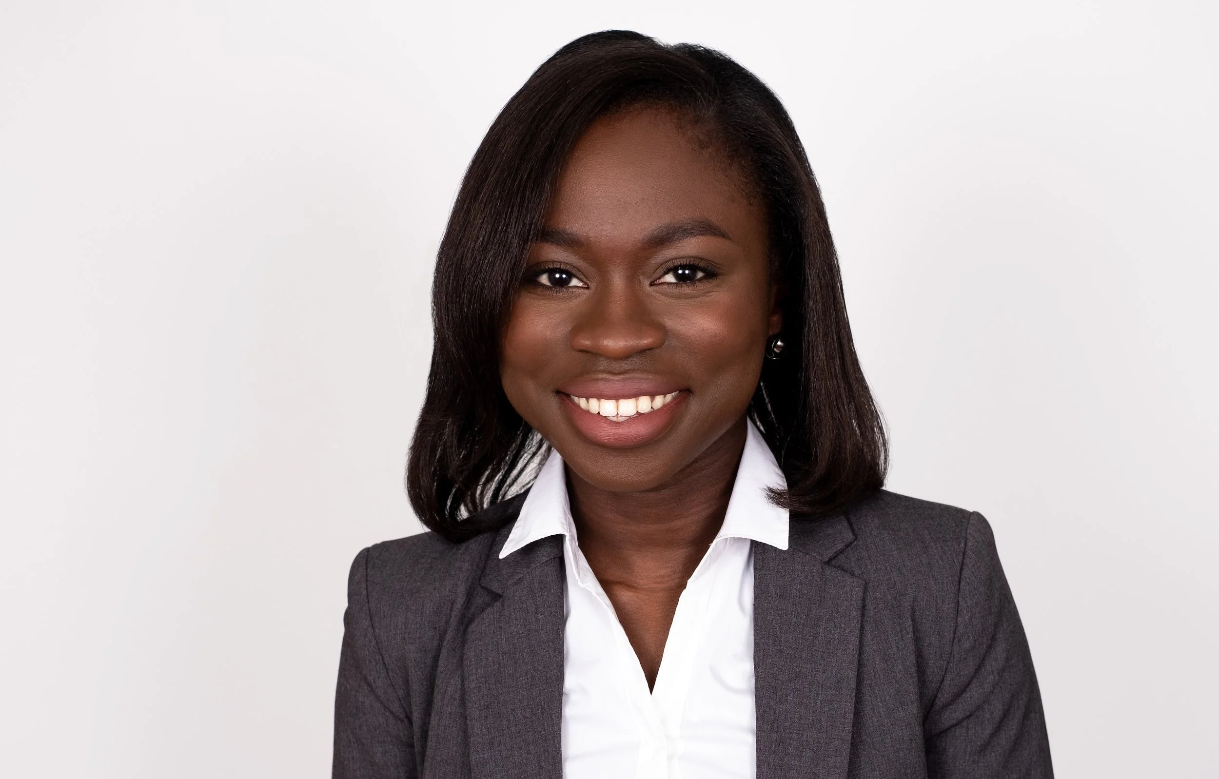 Professional Corporate Headshot by Twin Cities Photographer Jay Cupcake. Professional headshot of a smiling African American woman with shoulder-length dark hair, wearing a dark gray blazer and white collared shirt, against a plain light background.