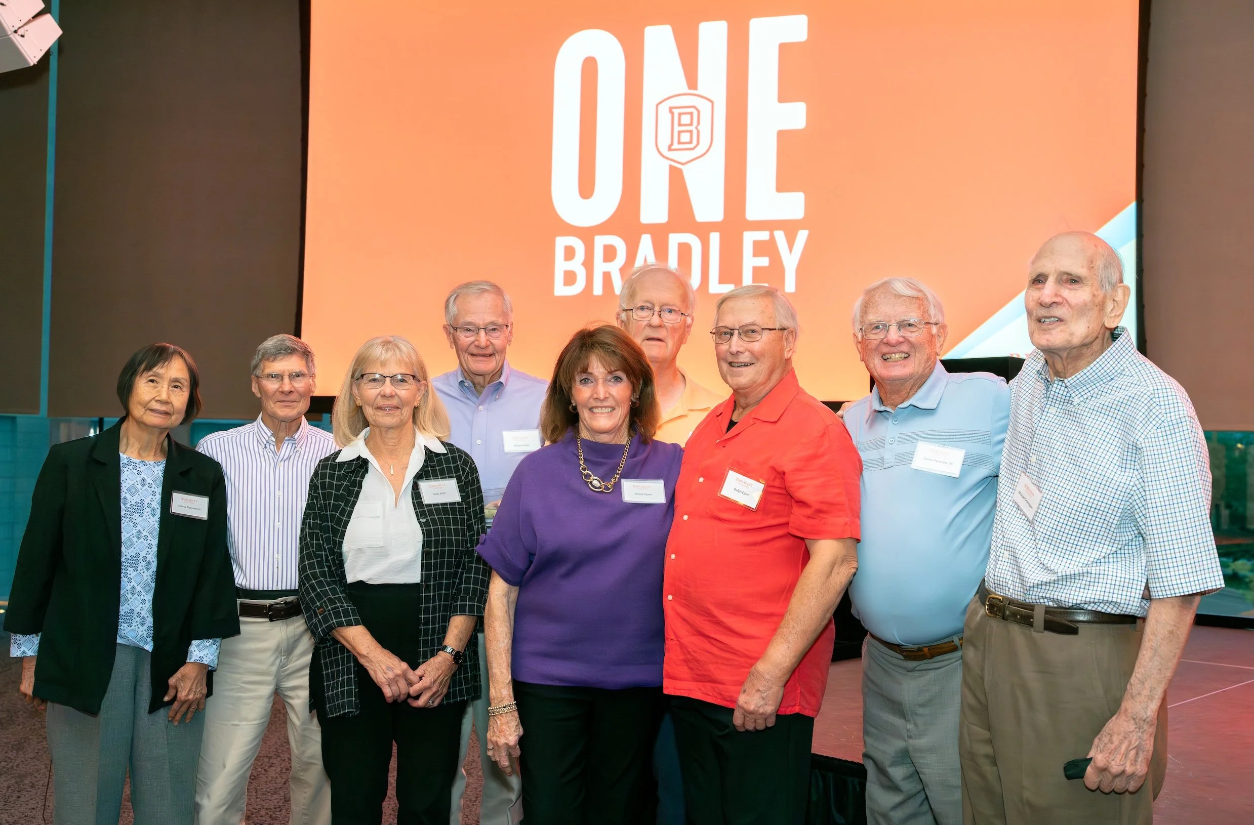 Professional Event Photography by Saint Paul Photographer Jay Cupcake. Group of nine older adults standing together at a conference or event, smiling for the camera, with a large screen behind them displaying 'One Bradley'.