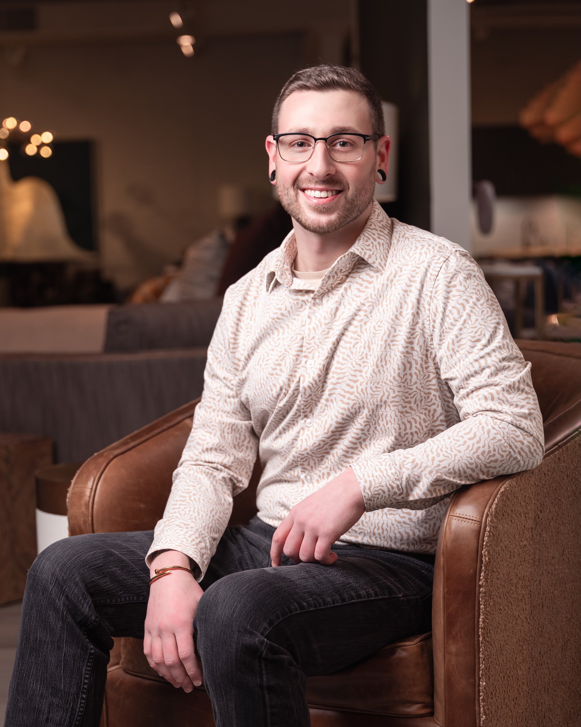 Professional Corporate Headshot by Saint Paul Photographer Jay Cupcake. A smiling man with short hair, glasses, and gauges in his ears, wearing a beige patterned shirt and black pants, sitting on a brown leather couch in a warmly lit room.