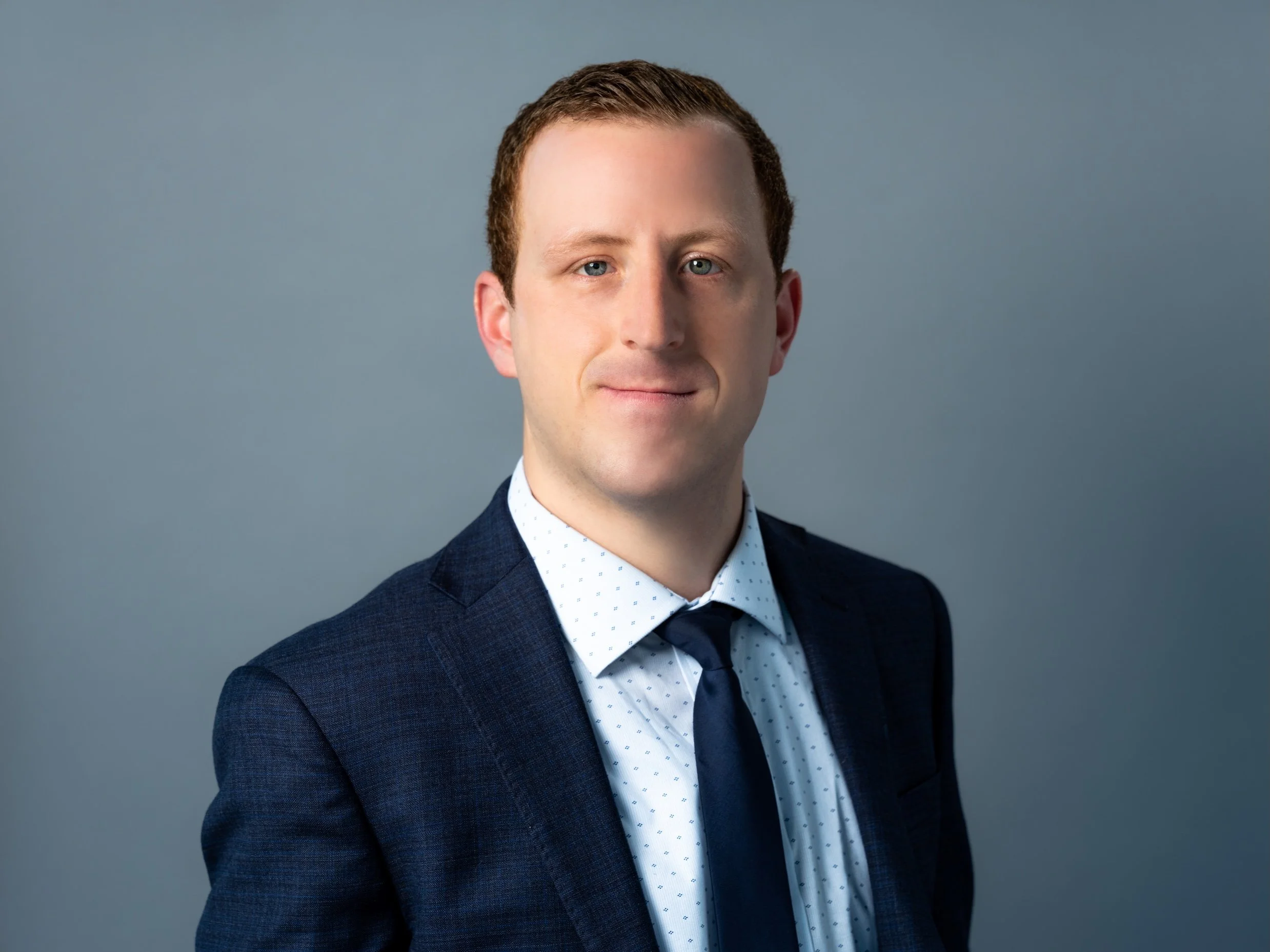 Professional Corporate Headshot by Saint Paul Photographer Jay Cupcake. Professional portrait of a man with short light brown hair, blue eyes, wearing a dark blue suit, white shirt, and navy tie, smiling subtly against a gray background.