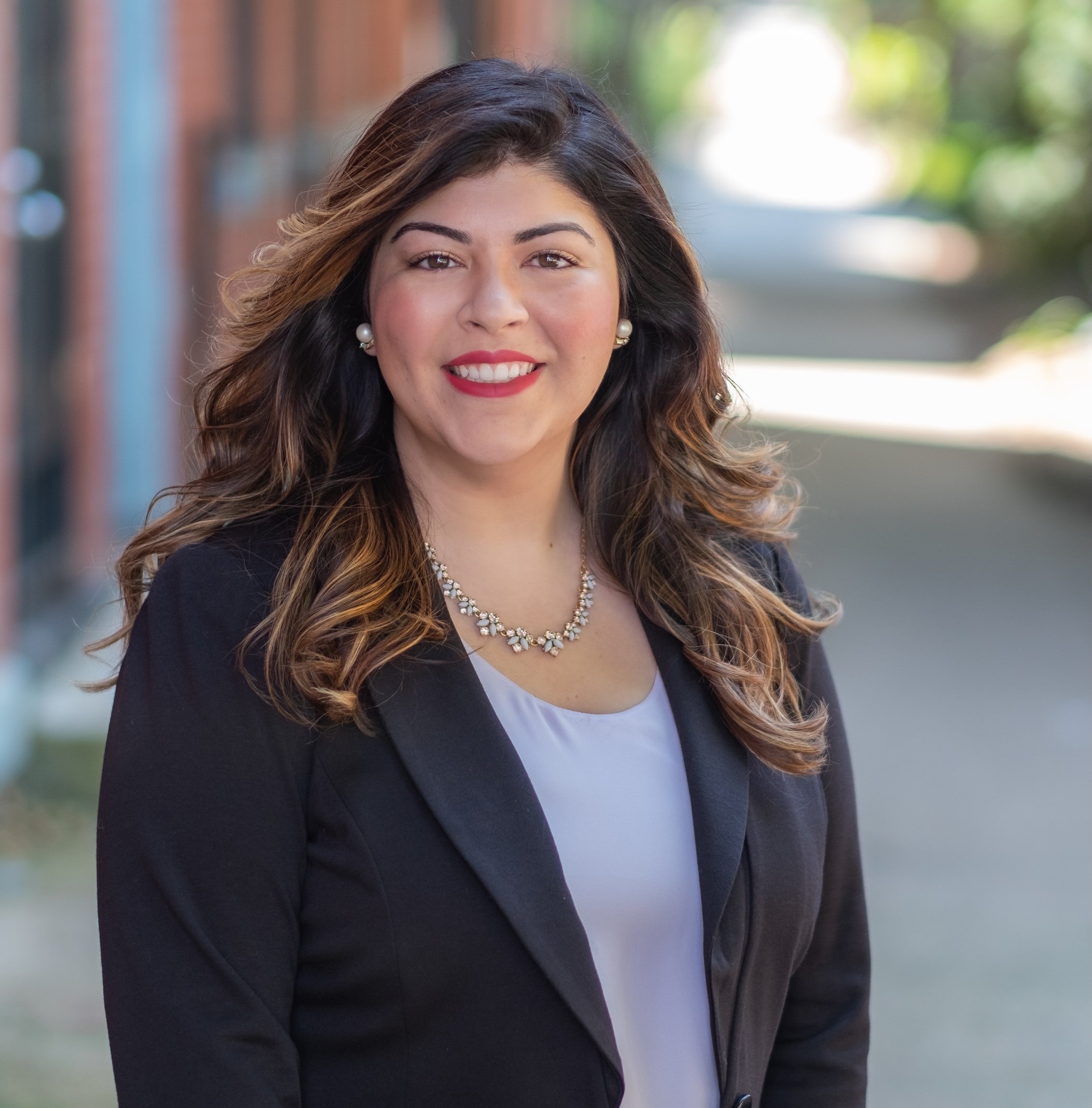xA professional woman with long, wavy brown hair, wearing a black blazer, white blouse, pearl earrings, and a pearl necklace, smiling outdoors with a blurred background of trees and buildings.
