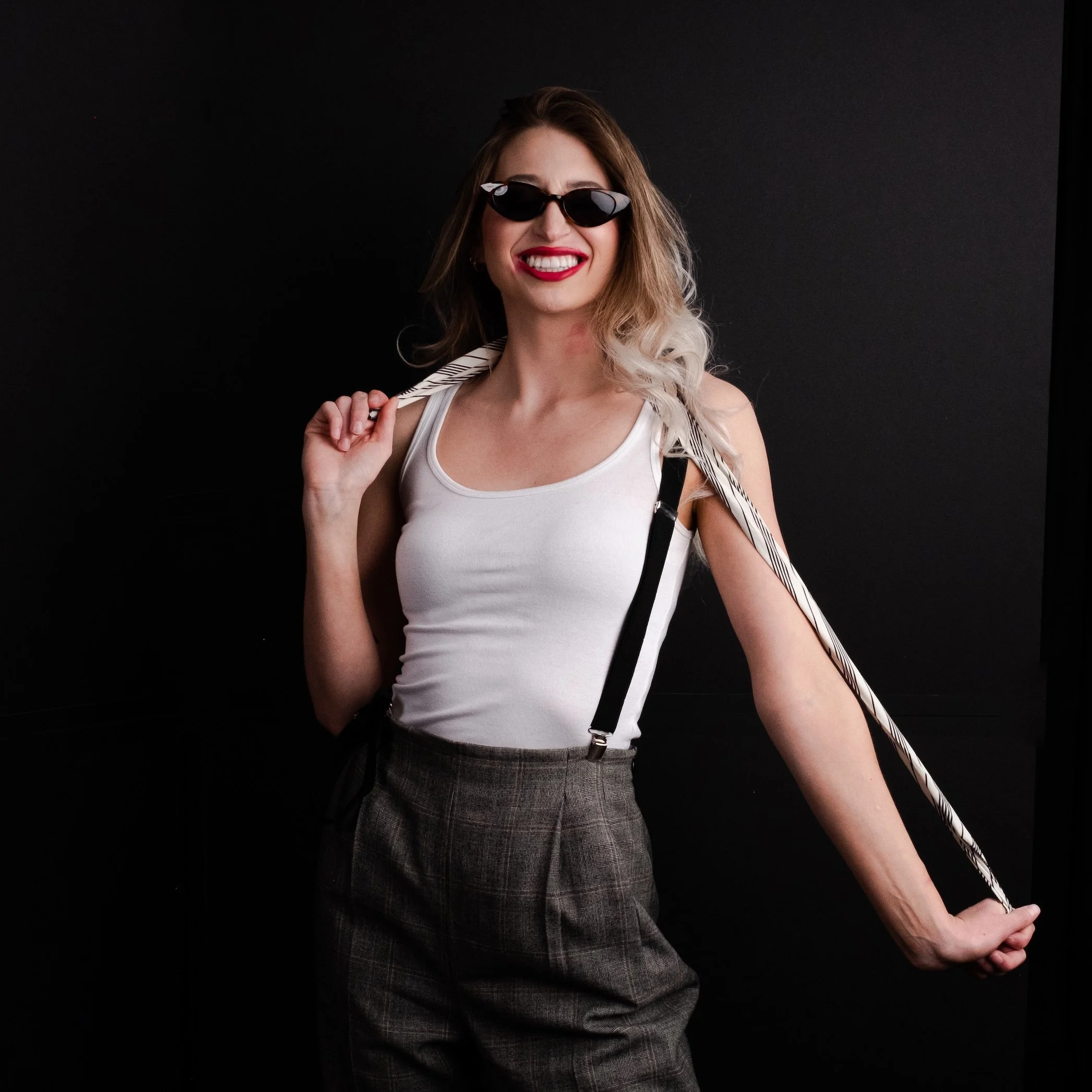 Professional Portrait Photography by Saint Paul Photographer Jay Cupcake. A woman with blonde and brown hair wearing sunglasses, a white tank top, high-waisted plaid pants, and holding a striped scarf over her shoulder, smiling against a black backgr