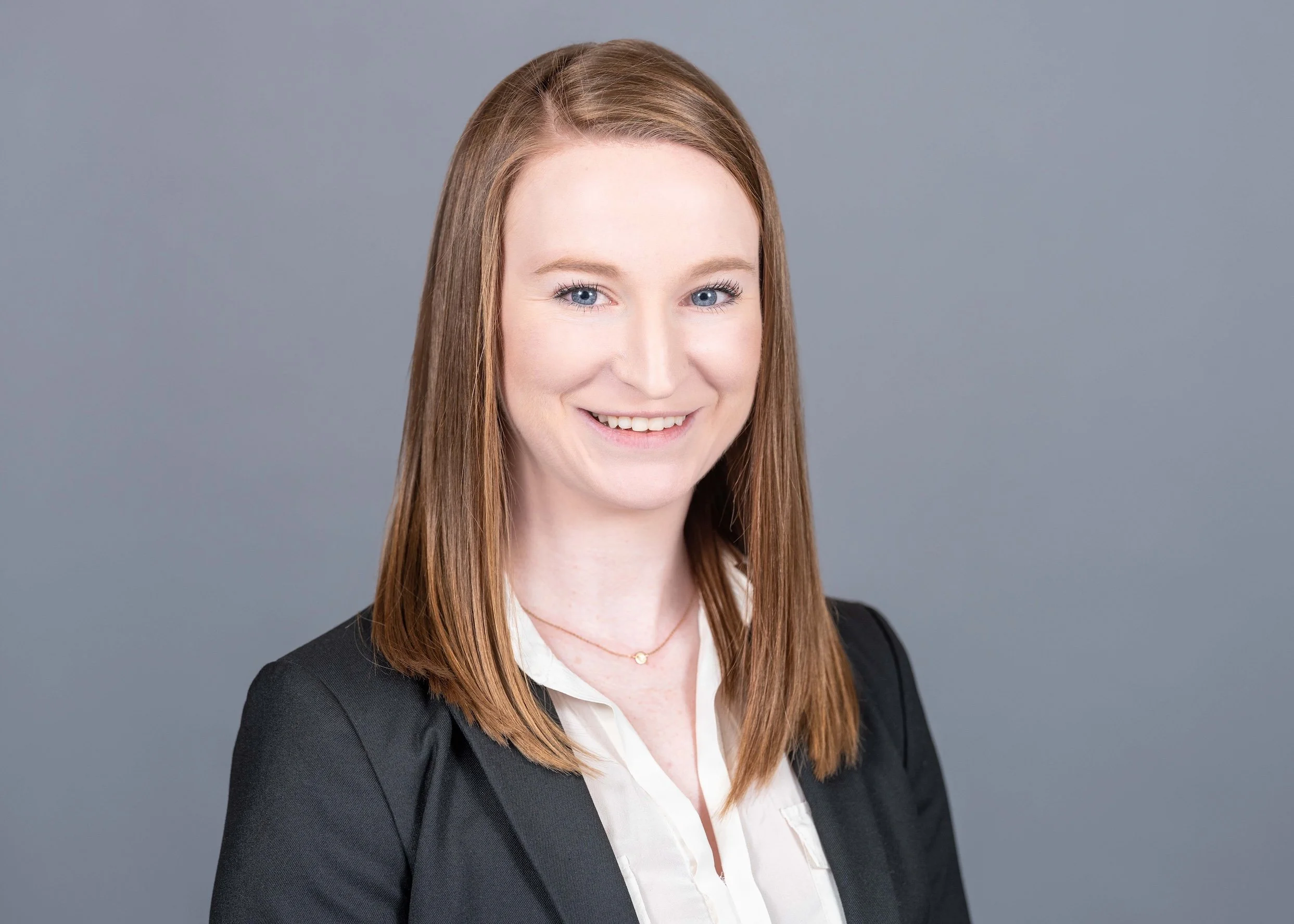 Professional Corporate Headshot by Minneapolis Photographer Jay Cupcake. A woman with shoulder-length red hair and a light skin tone, smiling, wearing a black blazer over a white blouse, against a plain gray background.