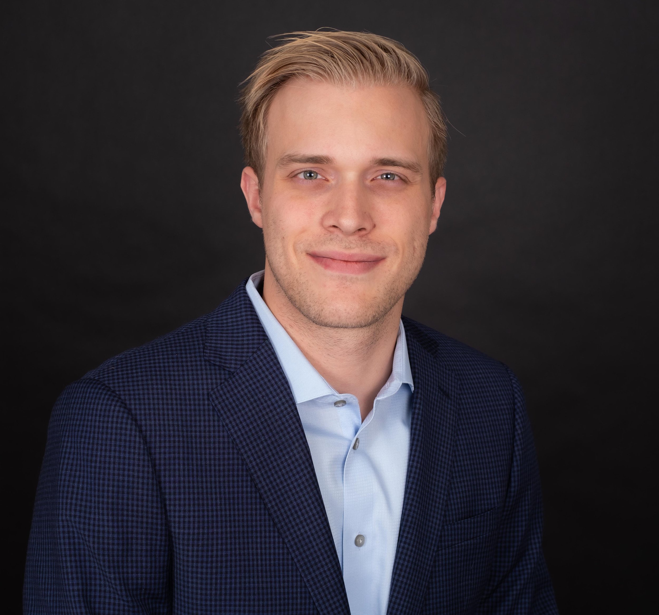 Professional Corporate Headshot by Minneapolis Photographer Jay Cupcake. A professional headshot of a young man with blond hair, wearing a blue checkered blazer and a light blue shirt, smiling gently against a black background.
