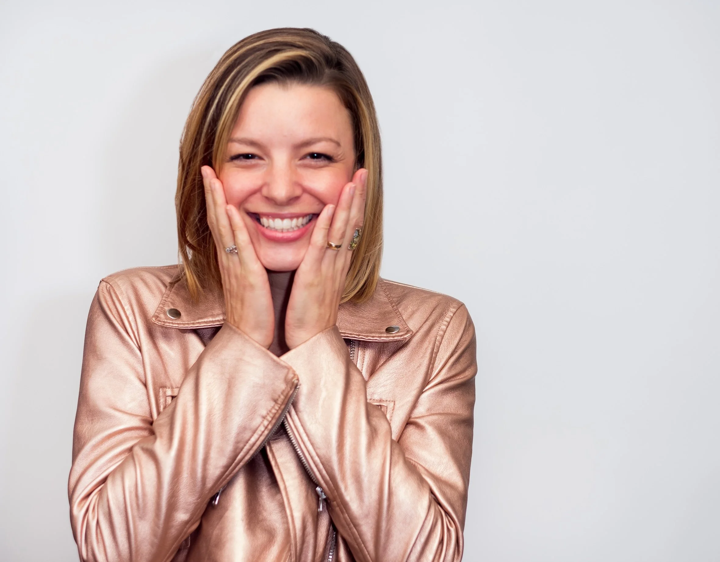 Professional Portrait Photography by Twin Cities Photographer Jay Cupcake. A woman with shoulder-length blonde hair smiling, wearing a shiny pinkish-beige leather jacket, touching her cheeks with both hands, standing against a plain white background.