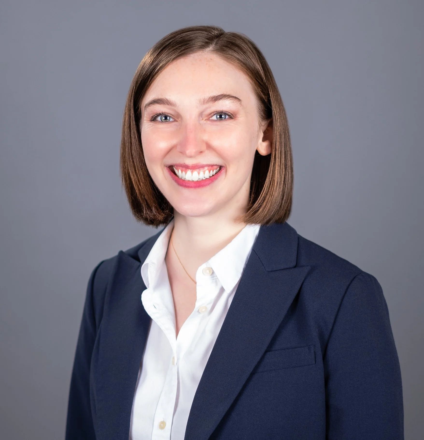 Professional Corporate Headshot by Twin Cities Photographer Jay Cupcake. Professional woman with a smile, wearing a dark blazer and white shirt, against a gray background.