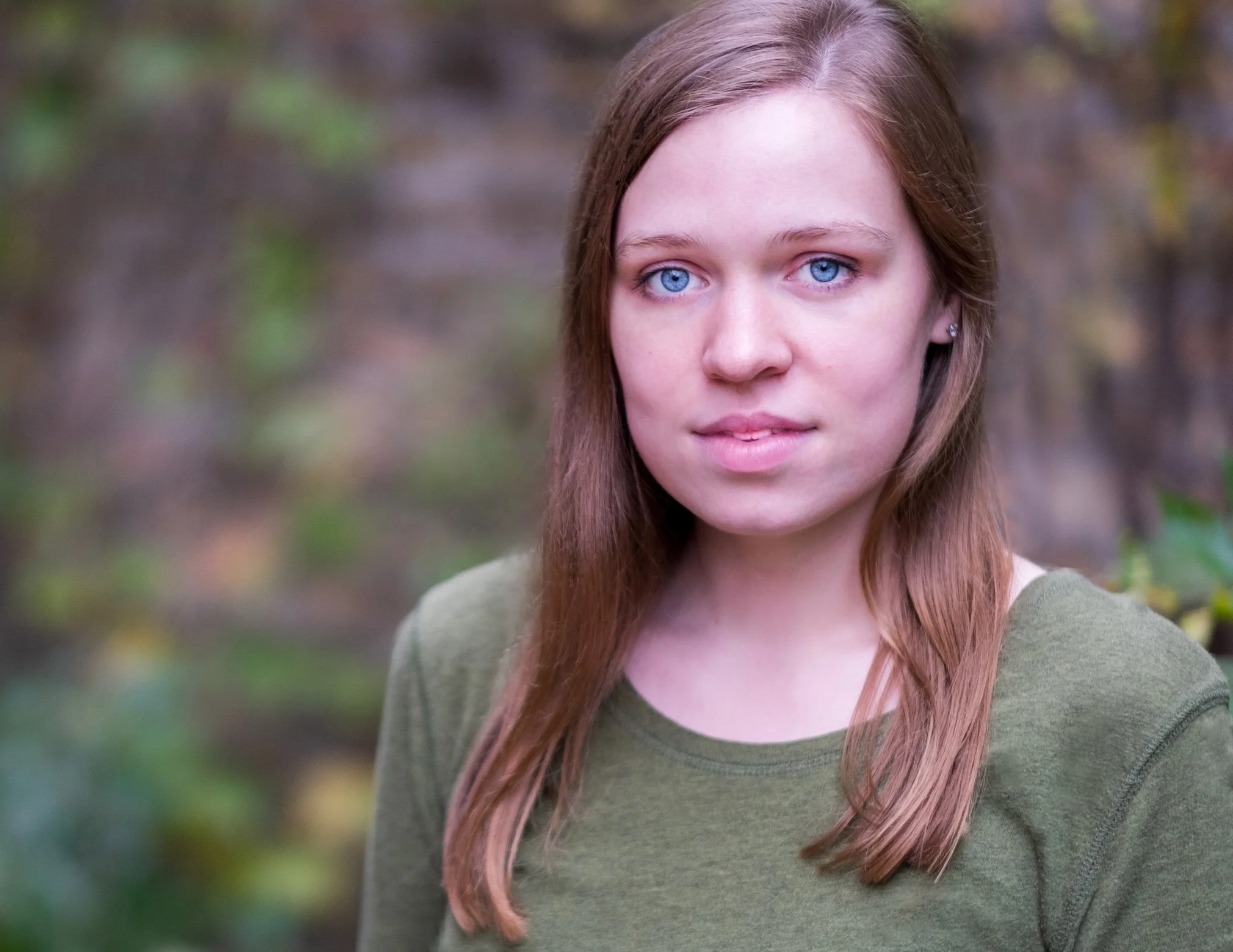 Professional Actor Headshot by Saint Paul Photographer Jay Cupcake. Young woman with long brown hair and blue eyes outdoors in a natural setting with blurred trees and foliage in the background.