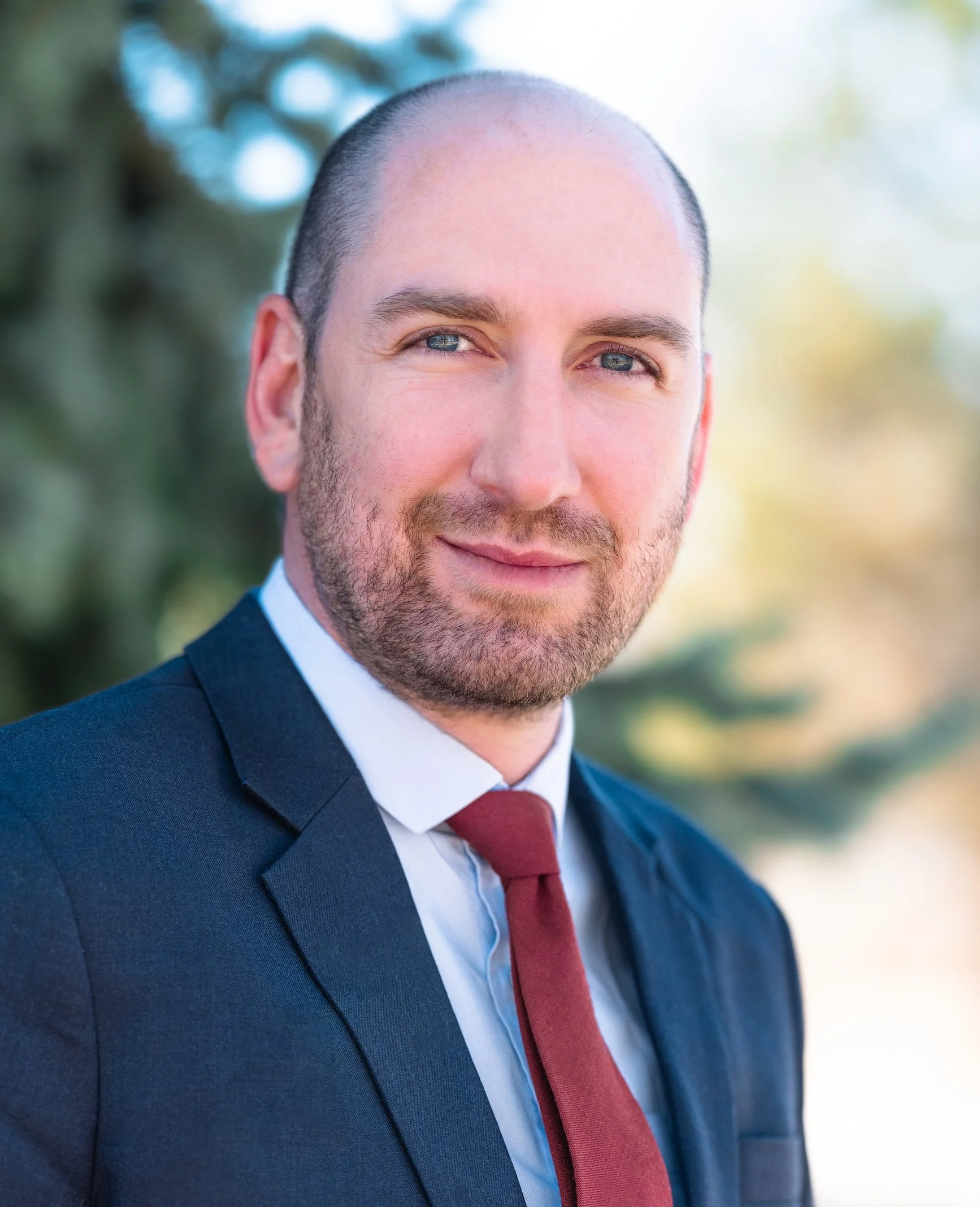Professional Corporate Headshot by Minneapolis Photographer Jay Cupcake. Portrait of a Caucasian man with a beard, wearing a dark suit, white shirt, and burgundy tie, outdoors with blurred green and yellow foliage in the background.