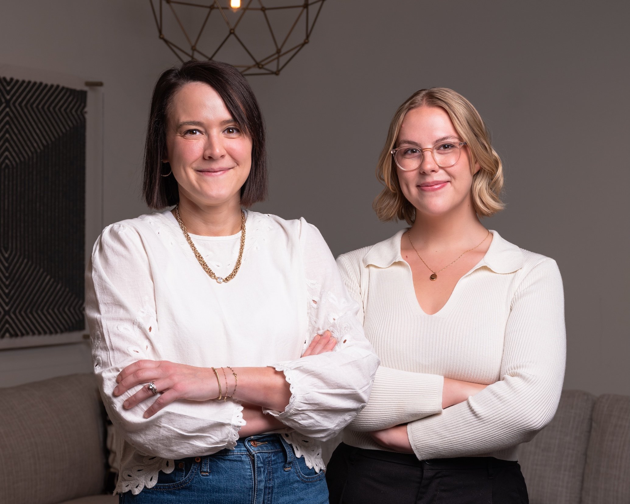 Professional Corporate Headshot by Twin Cities Photographer Jay Cupcake. Two women standing side by side indoors with their arms crossed, smiling at the camera. The woman on the left has dark shoulder-length hair, wearing a white blouse with eyelet d