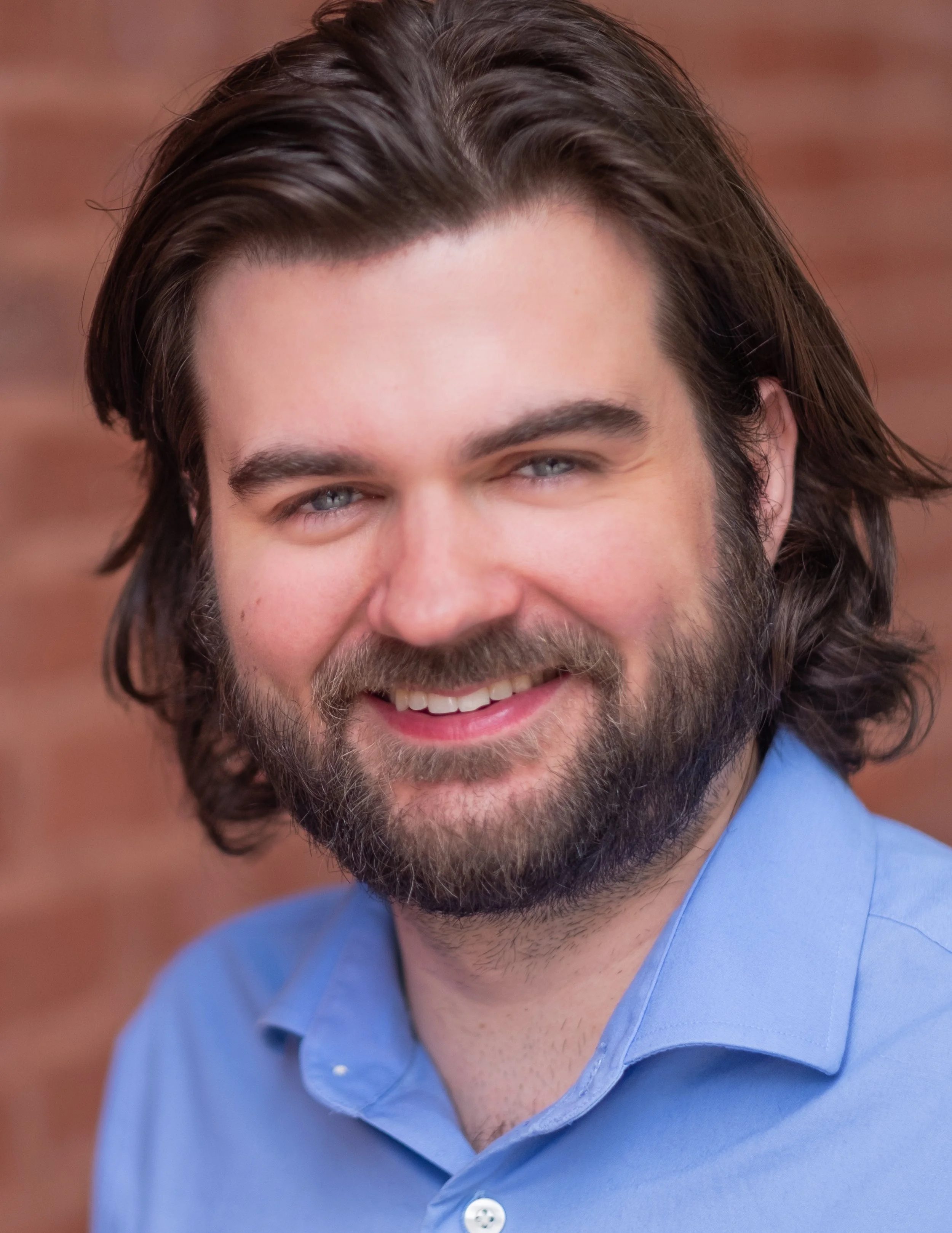 Professional headshot by St. Paul photographer Jay Cupcake. A smiling man with long brown hair, a beard, and blue eyes, wearing a light blue collared shirt, against a blurred brick wall background.
