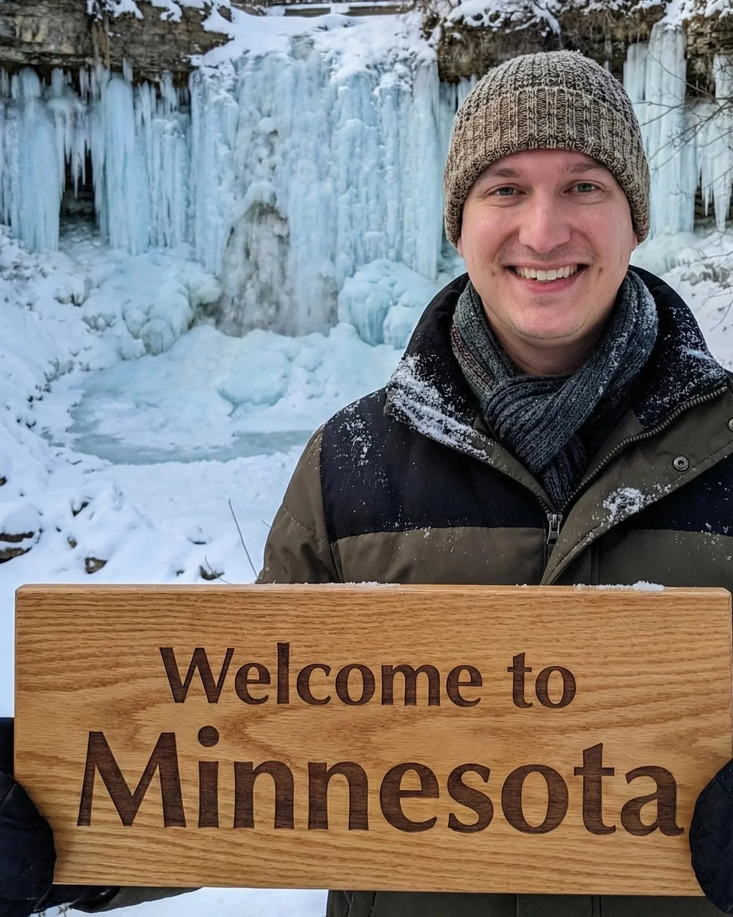 A smiling man holding a wooden sign that says 'Welcome to Minnesota' with a snowy winter landscape and an ice-covered waterfall in the background.