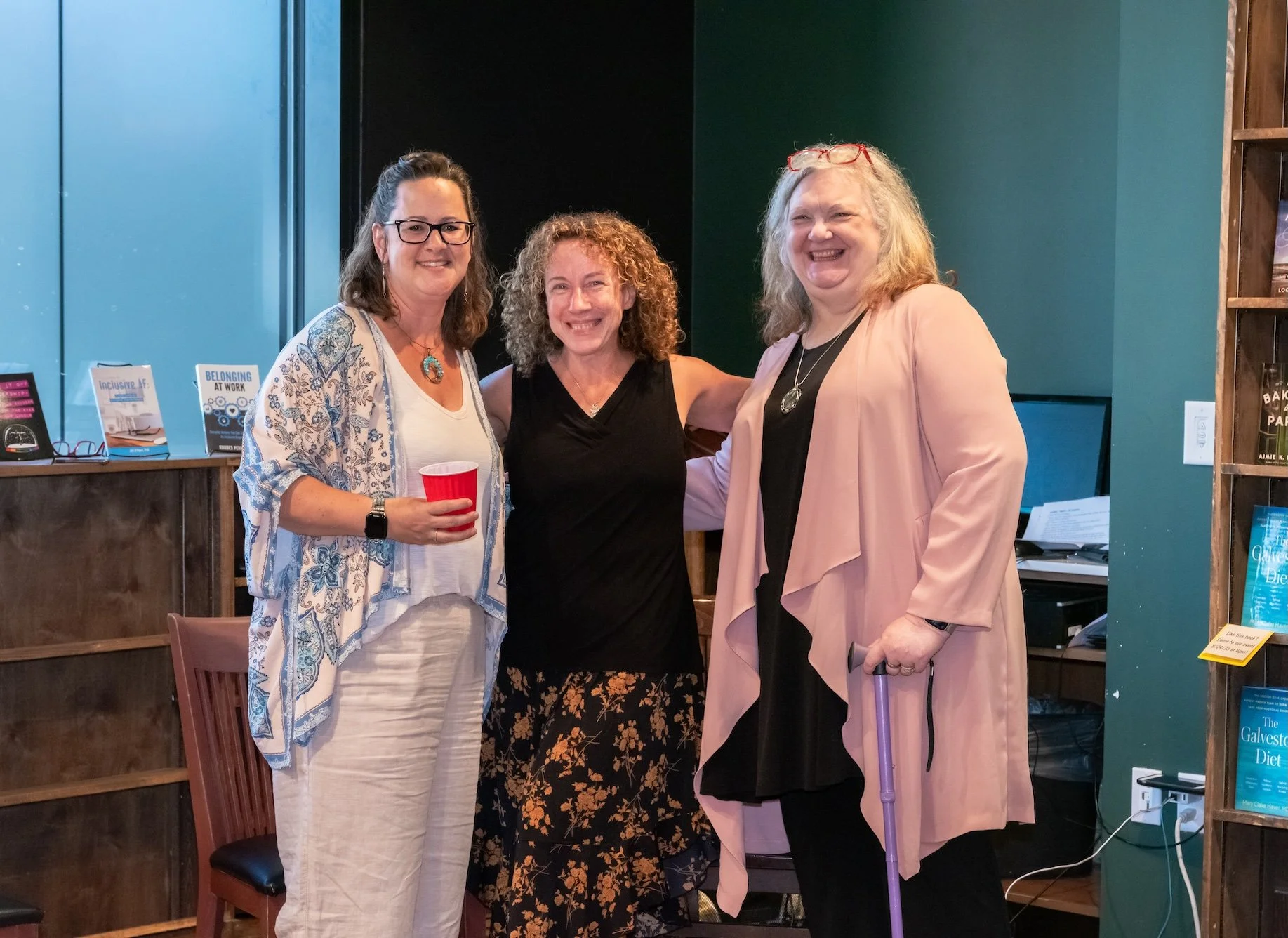 Professional Event Photography by Twin Cities Photographer Jay Cupcake. Three women posing together, smiling, indoors at a social event with books and pamphlets in the background.