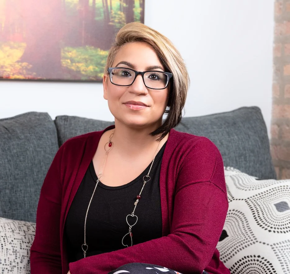 Professional Corporate Headshot by Minneapolis Photographer Jay Cupcake. A woman with short, blonde and brown hair, wearing glasses, a black top, a red cardigan, and a necklace, sitting on a gray couch with patterned pillows in a living room with a p