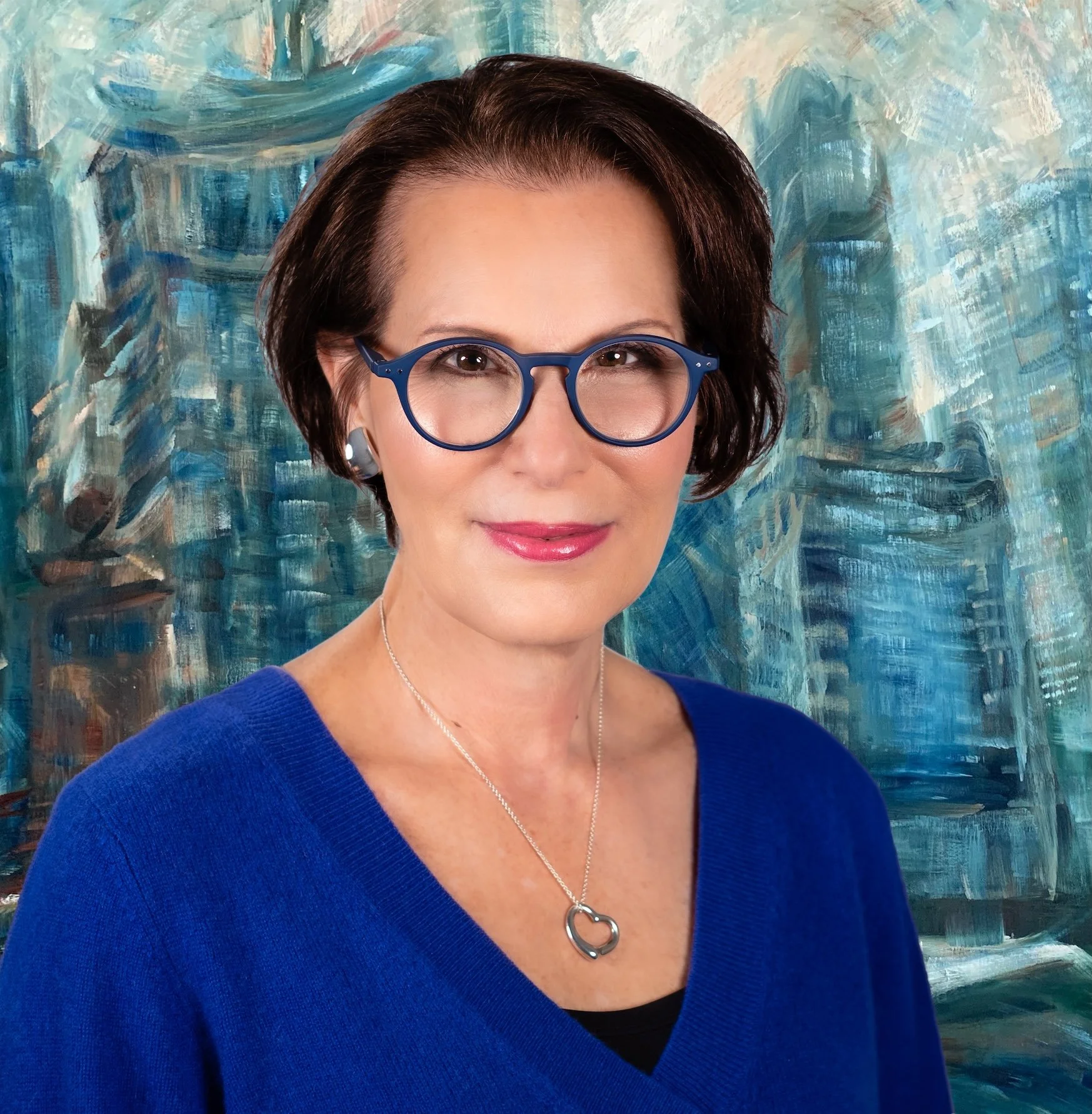 Professional Corporate Headshot by Twin Cities Photographer Jay Cupcake. A woman with short brown hair wearing blue glasses, a blue sweater, a silver necklace with a heart pendant, and earrings, standing in front of an abstract blue and teal backgrou