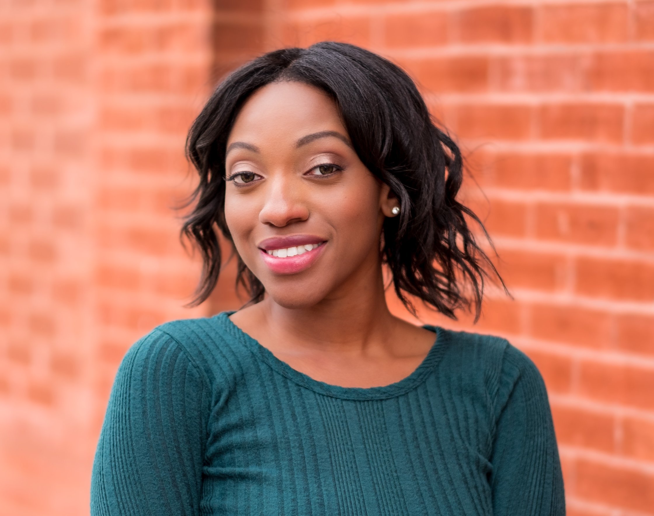 Professional Actor Headshot by Minneapolis Photographer Jay Cupcake. A smiling woman with shoulder-length curly black hair, wearing a teal long-sleeve top, standing in front of a blurred brick wall.