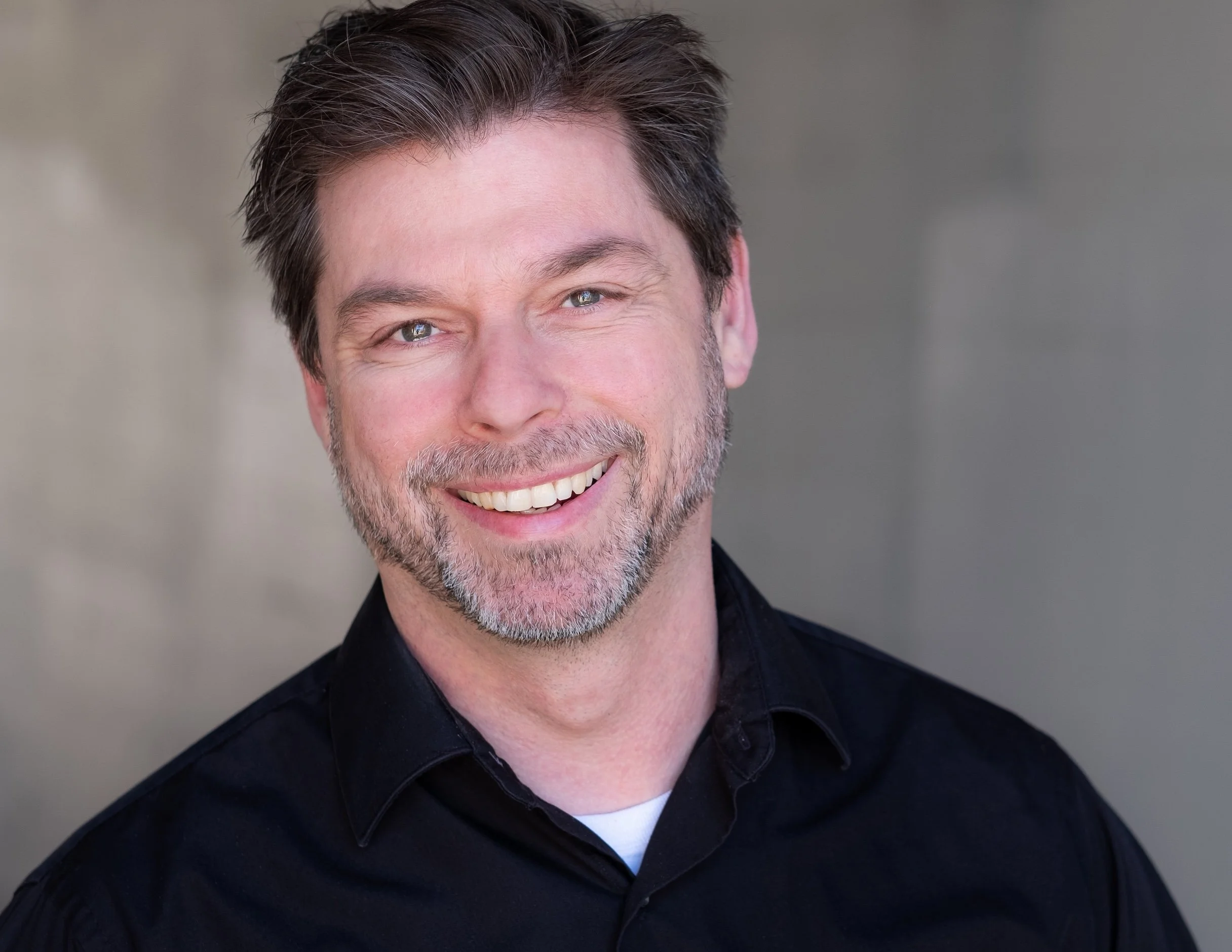 Professional Actor Headshot by Saint Paul Photographer Jay Cupcake. Headshot of a smiling man with dark hair, a beard, and blue eyes, wearing a black collared shirt, against a neutral background.