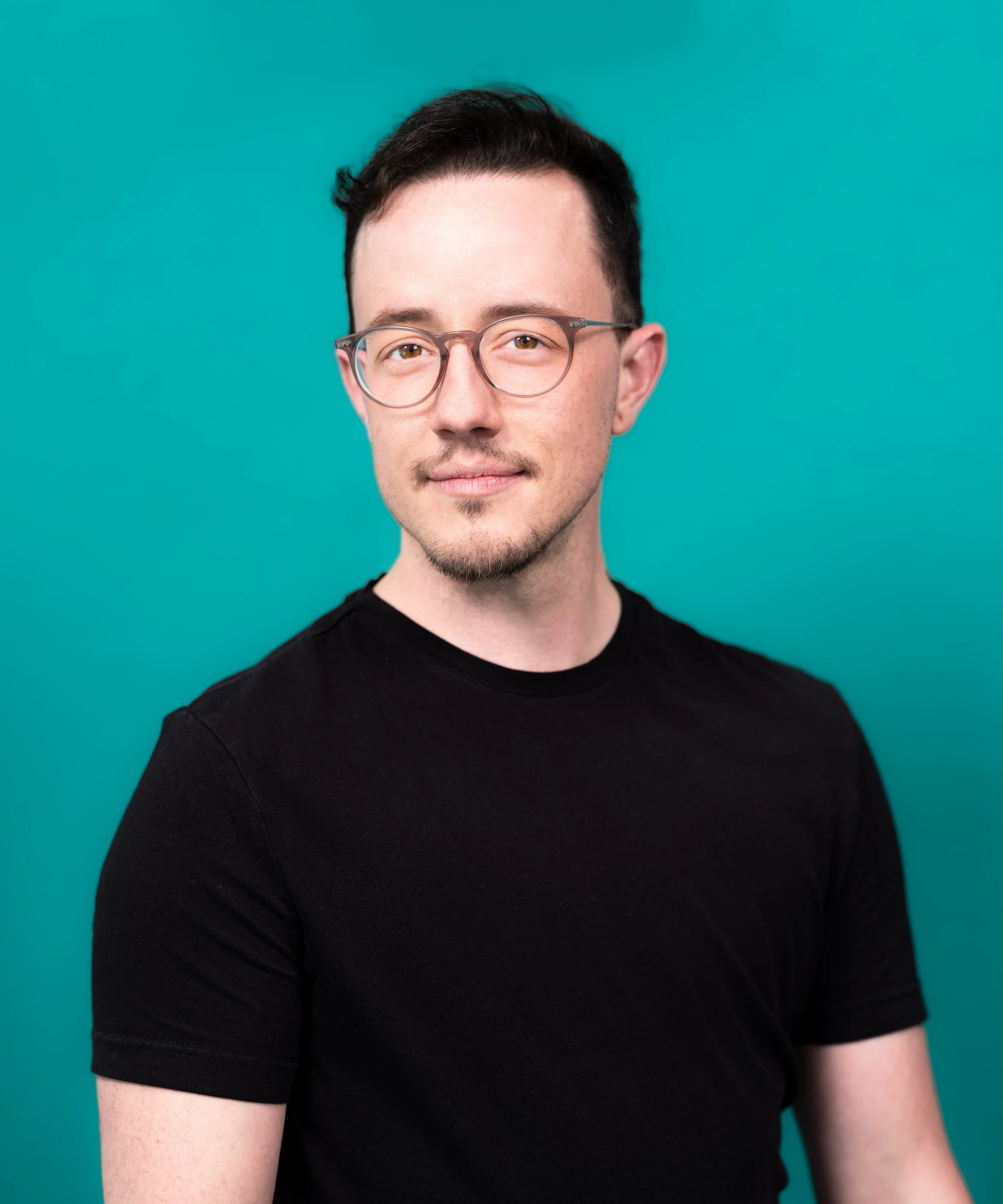 Professional Portrait Photography by Twin Cities Photographer Jay Cupcake. Portrait of a young man with glasses and dark hair, wearing a black t-shirt, standing against a teal background.