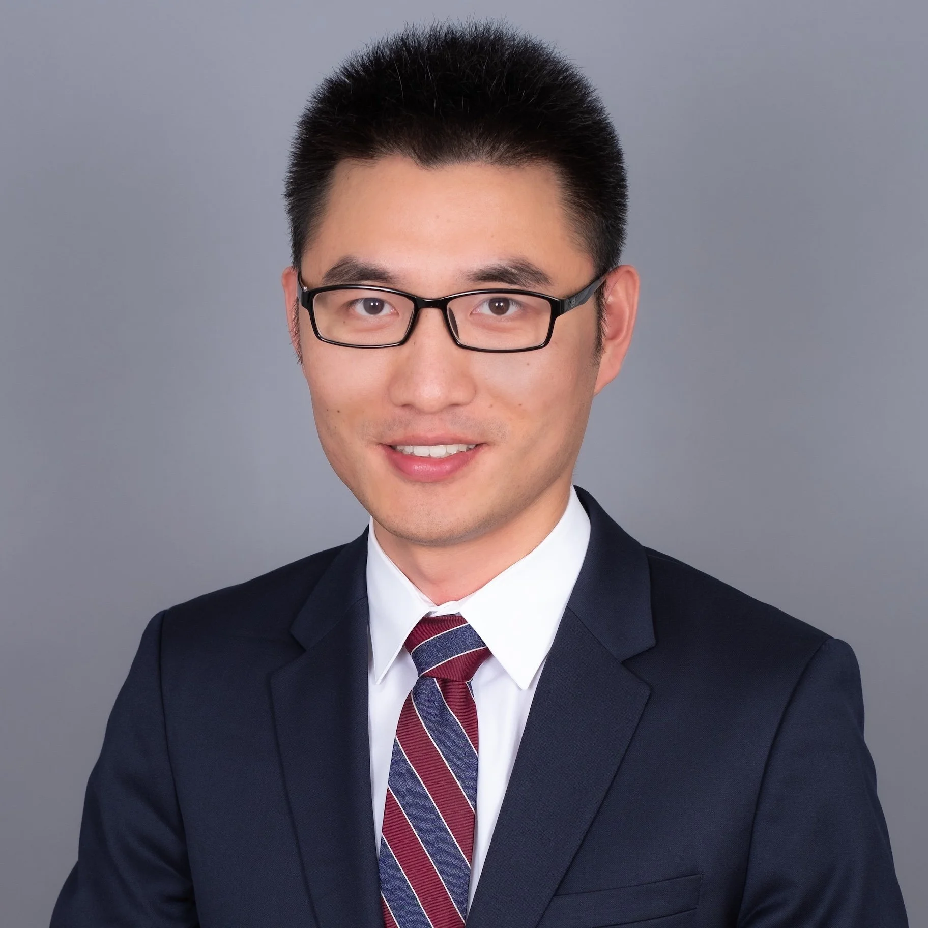 Professional Corporate Headshot by Saint Paul Photographer Jay Cupcake. Professional portrait of a young man with glasses wearing a dark suit, white shirt, and striped maroon and blue tie, smiling against a plain gray background.
