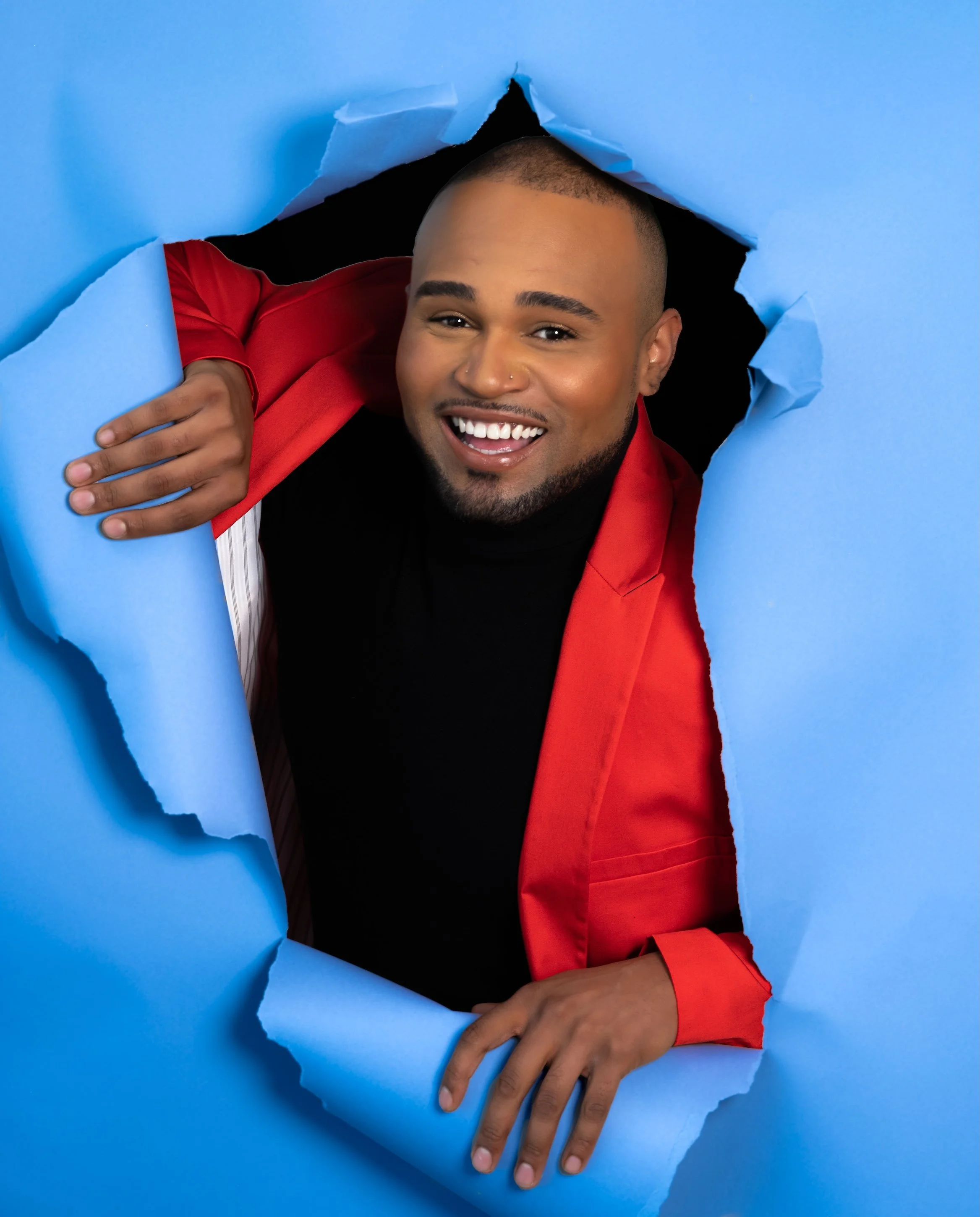Professional Portrait Photography by Saint Paul Photographer Jay Cupcake. A smiling man with a beard and short hair poking through a torn blue paper background, wearing a red blazer over a black shirt.