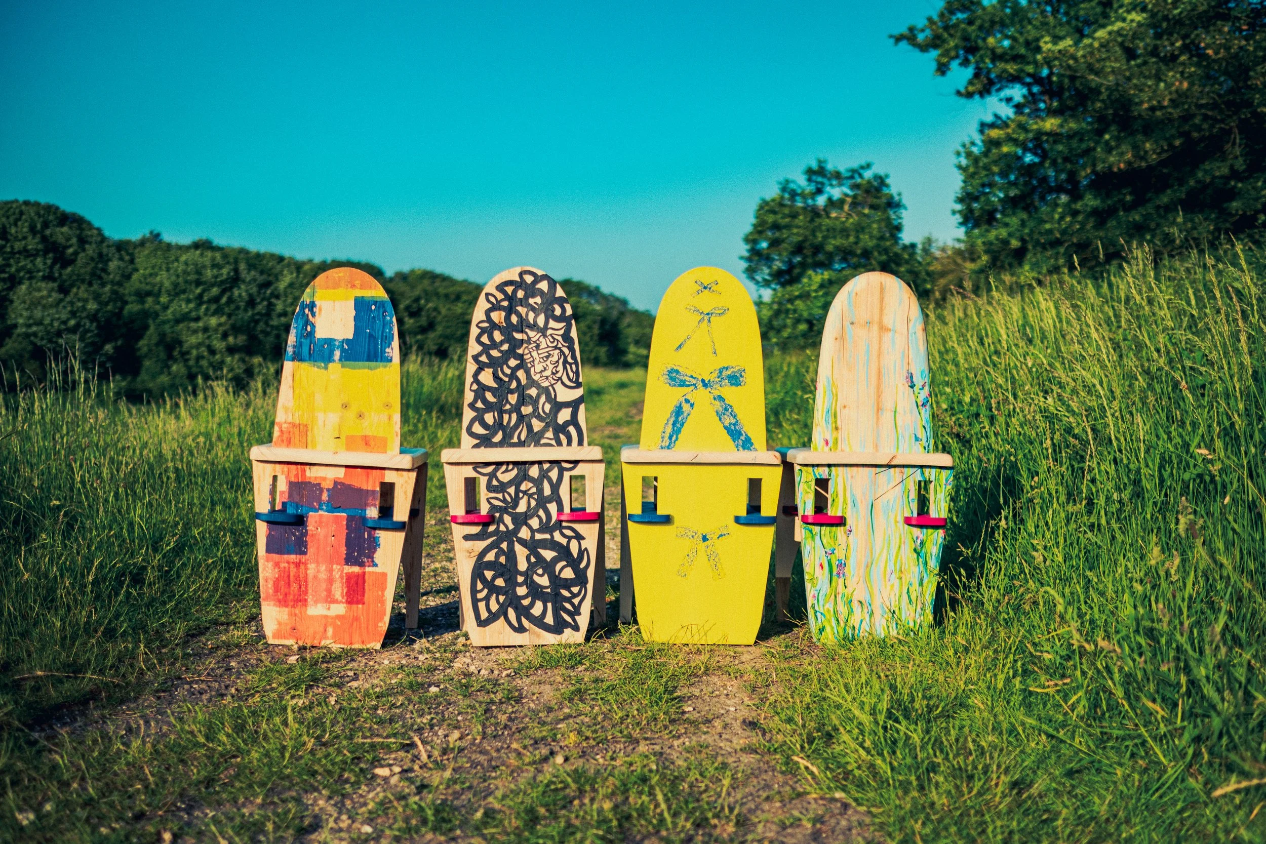 Four colorful painted wooden chairs on a grassy trail in a natural outdoor setting with trees and a bright blue sky. Photographed by Owen Mills
