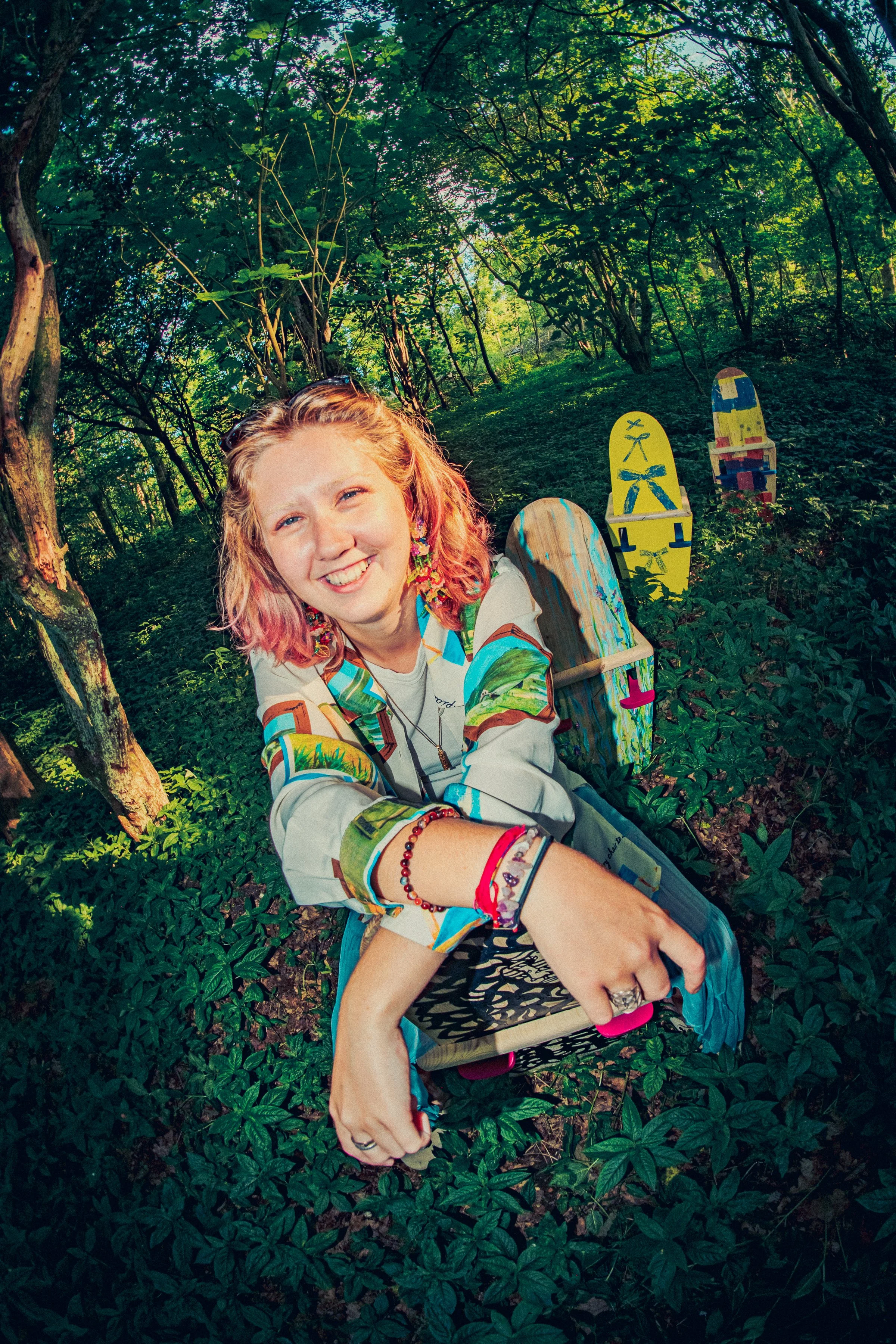 A young woman with pink and blonde hair smiling and taking a selfie in a forest with green trees and foliage, sitting on a wooden chair with colorful painted chairs in the background. @katyawaltonstudio instagram post photographed by Owen mills