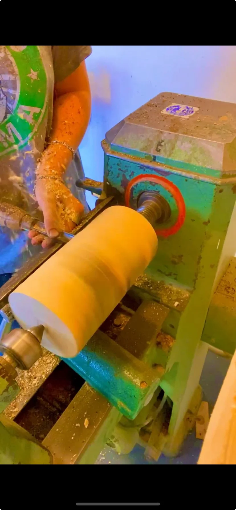 A person operating a green and orange woodworking lathe machine shaping a large cylindrical piece of wood. @katyawaltonstudio instagram post