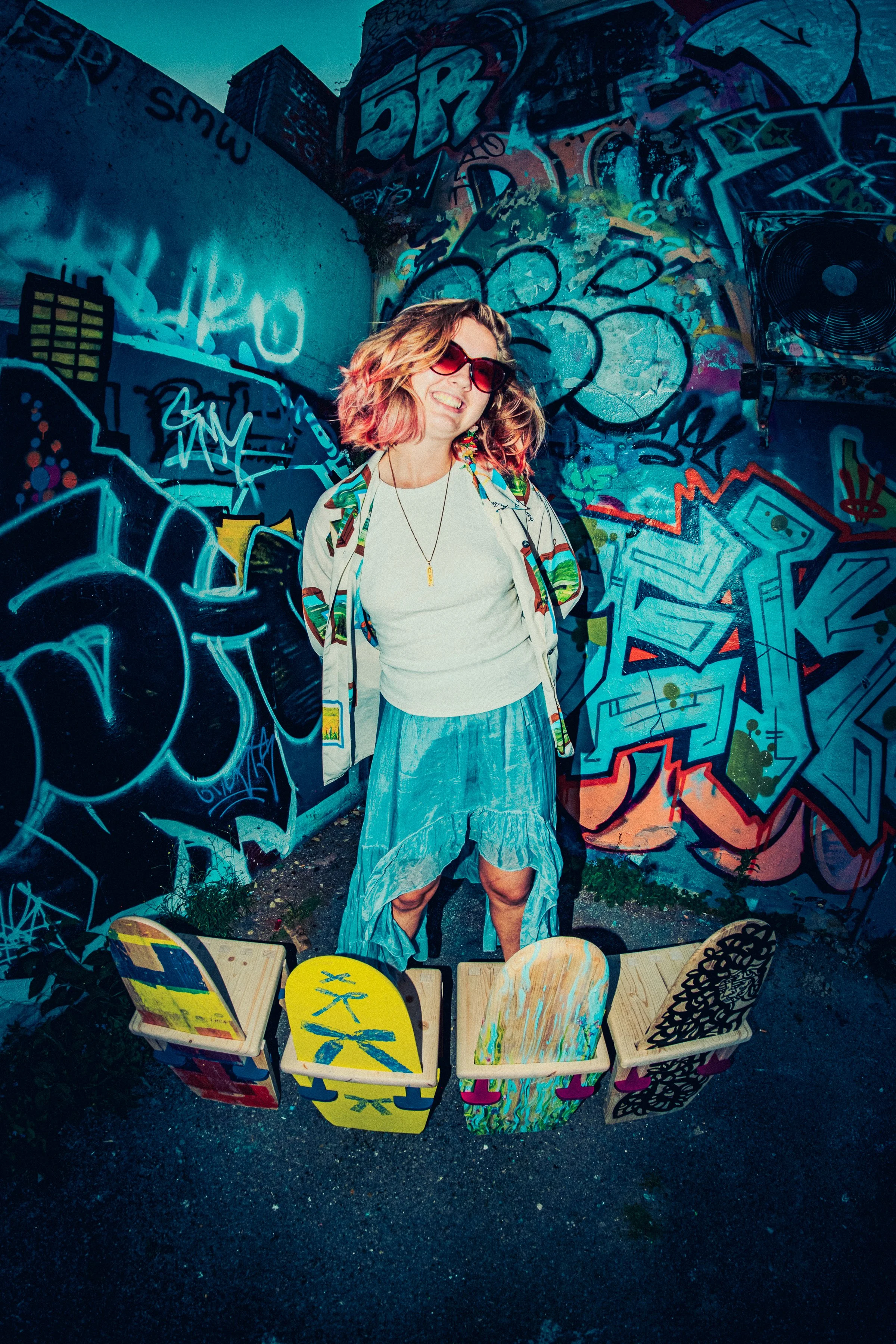 A woman with curly, pinkish hair, wearing sunglasses, standing in an alley surrounded by vibrant graffiti. There are painted chairs on the ground in front of her. Photographed by Owen Mills - Brighton