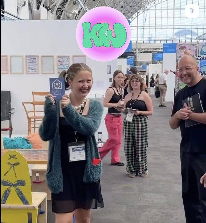 Group of people at a the New Designers trade show or exhibition, Andrew Tanner, awarding a recognition prize from Habitat UK , with booths and artwork in the background.