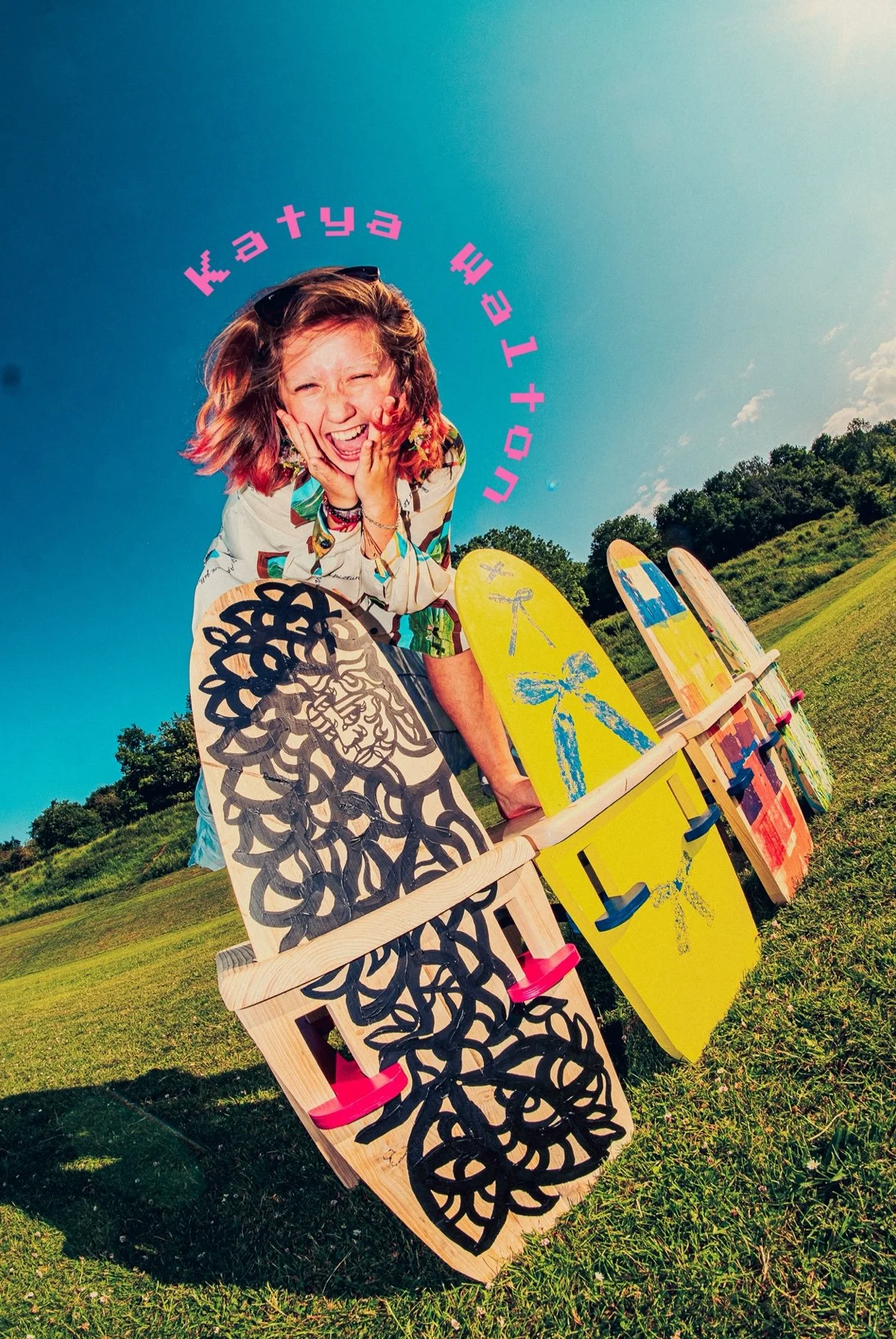 A young woman with colorful hair and a joyful expression sits outside on a sunny day, surrounded by painted wooden handmade chairs arranged in a semi-circle on the grassy field. @katyawaltonstudio instagram post Owen mills Brighton
