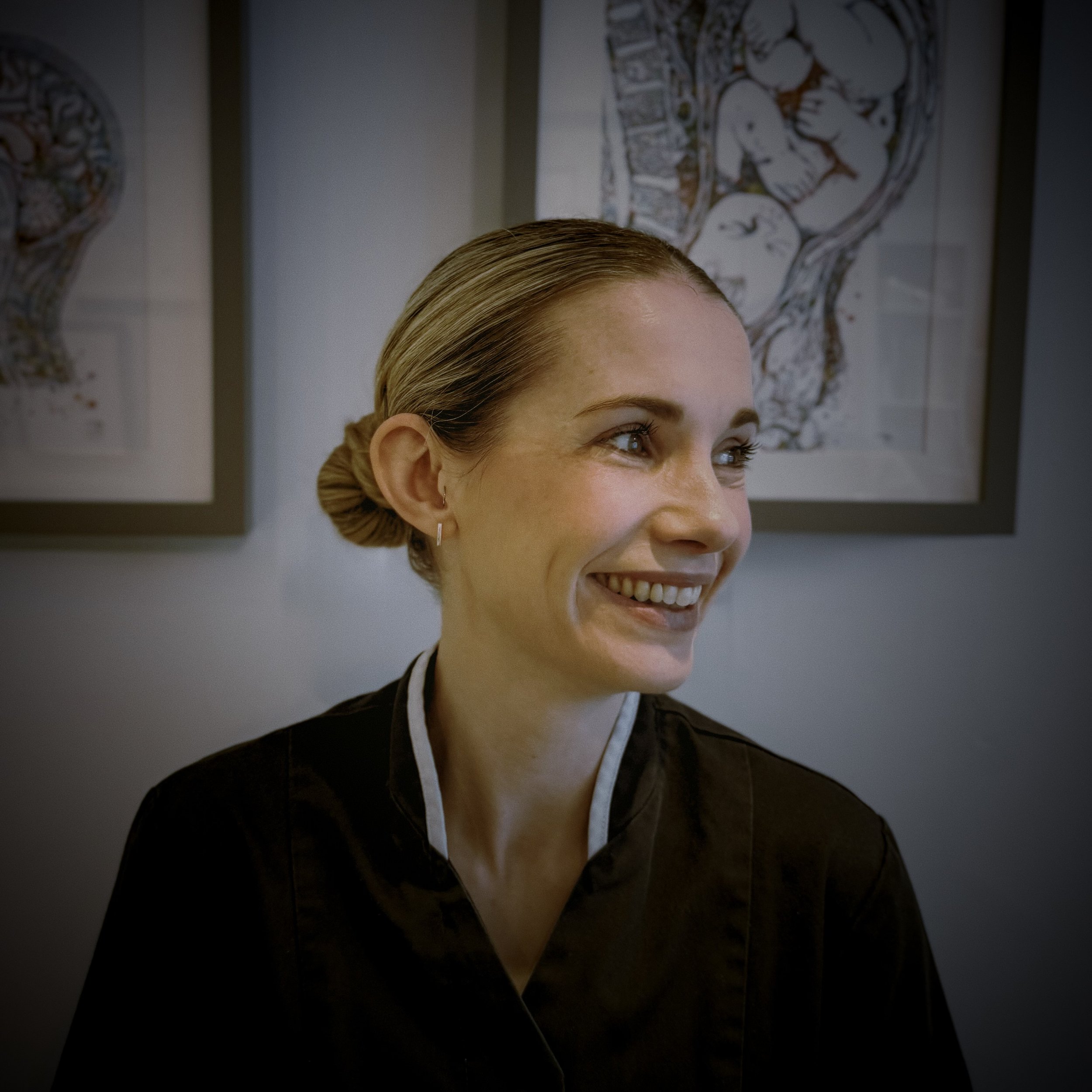 Close-up of a woman with blond hair styled in a bun, smiling, with artwork featuring human figures in the background.