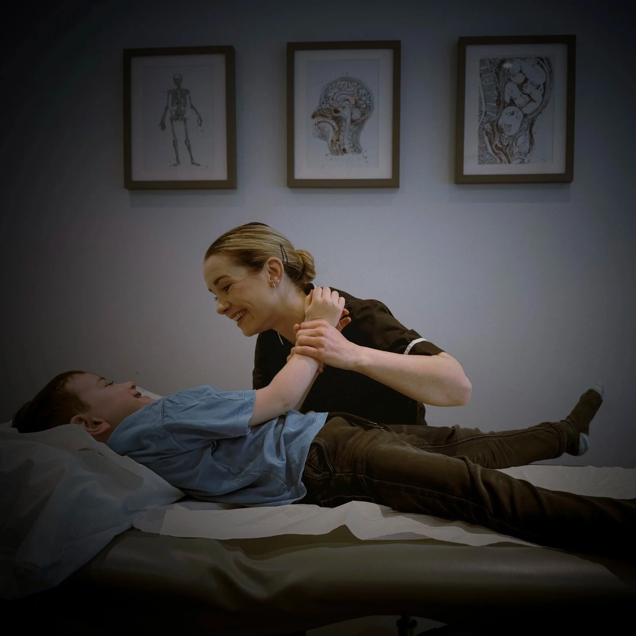 A woman leaning over a hospital bed and smiling at a young boy lying in bed, holding his hands. The background features framed medical illustrations on the wall.