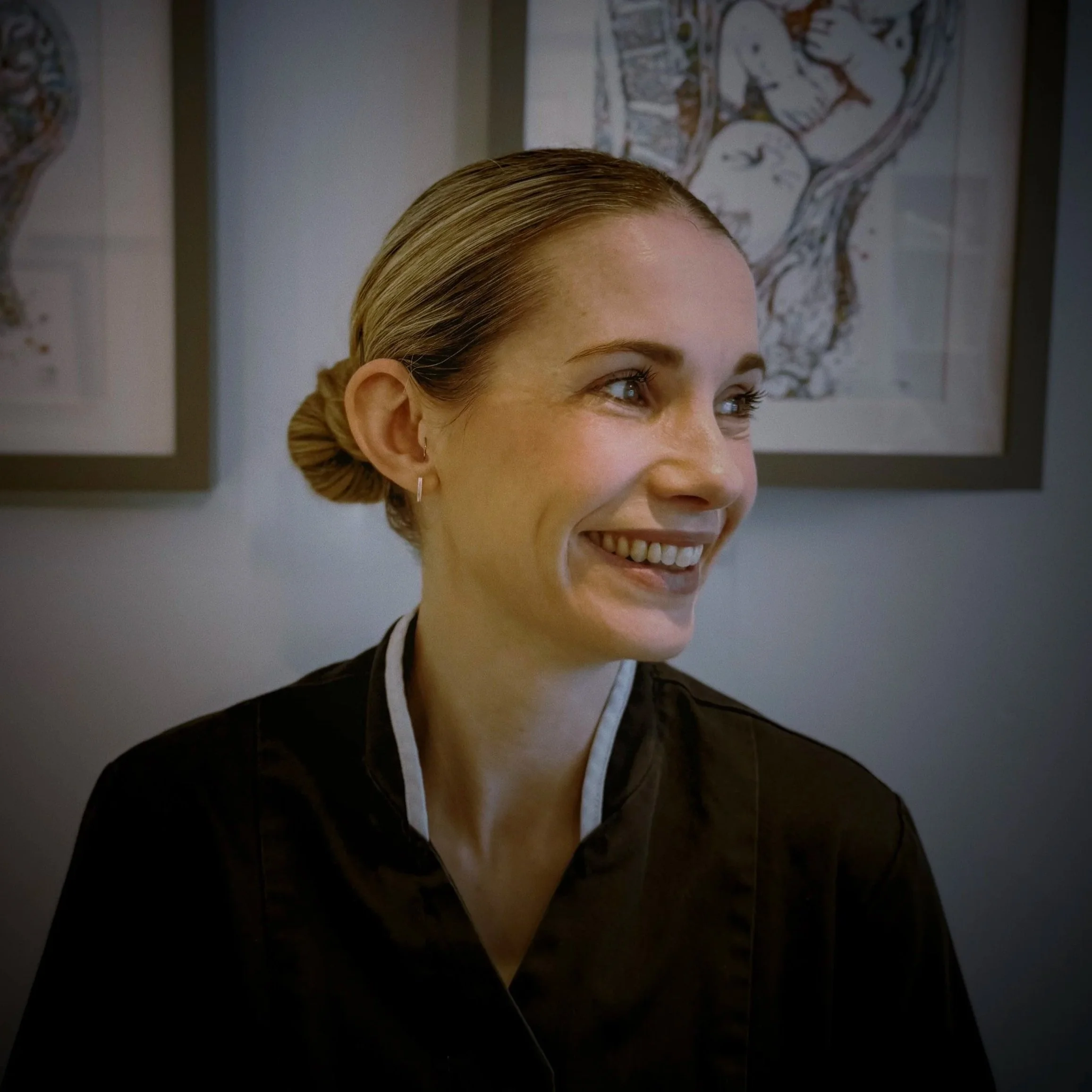 Close-up of a smiling woman with blonde hair styled in a bun, wearing earrings and a dark outfit, with artwork hanging on the wall in the background.
