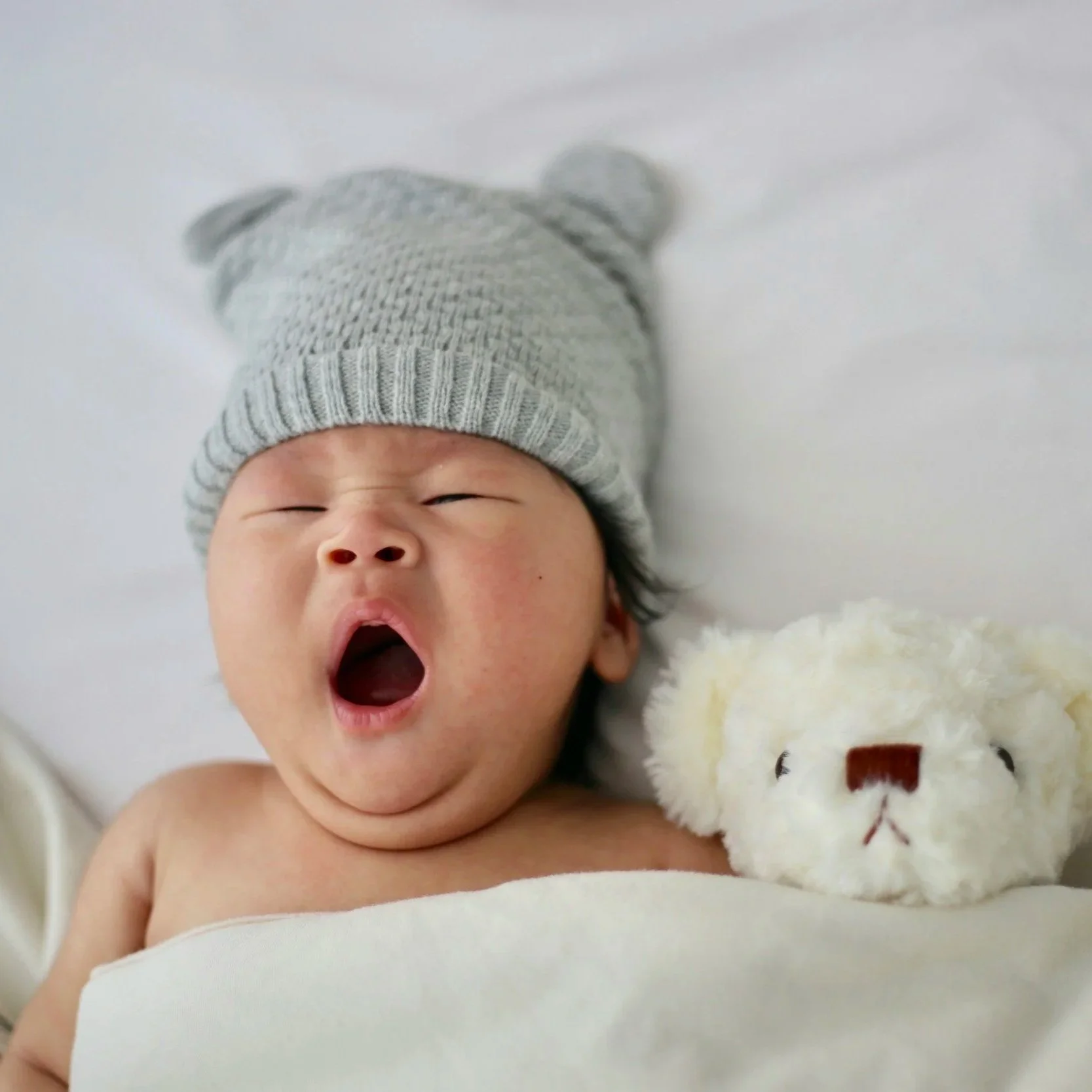A yawning baby with a grey knit hat, lying on a white blanket with a white teddy bear nearby.