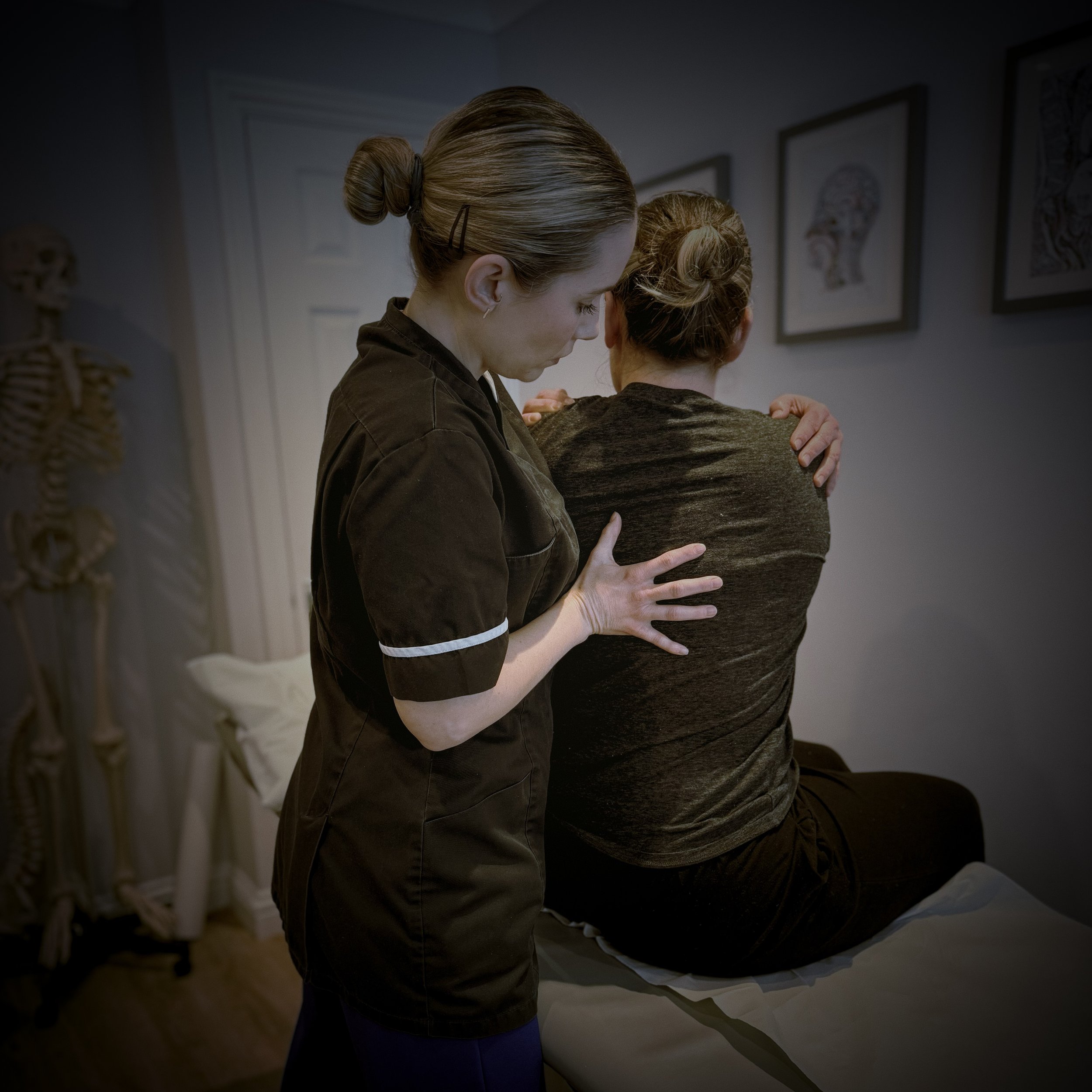 Two women embrace in an art classroom, one facing the wall with framed artwork and anatomical drawings, the other with hair in a bun, wearing a black shirt, hugging the other woman who is also in black.