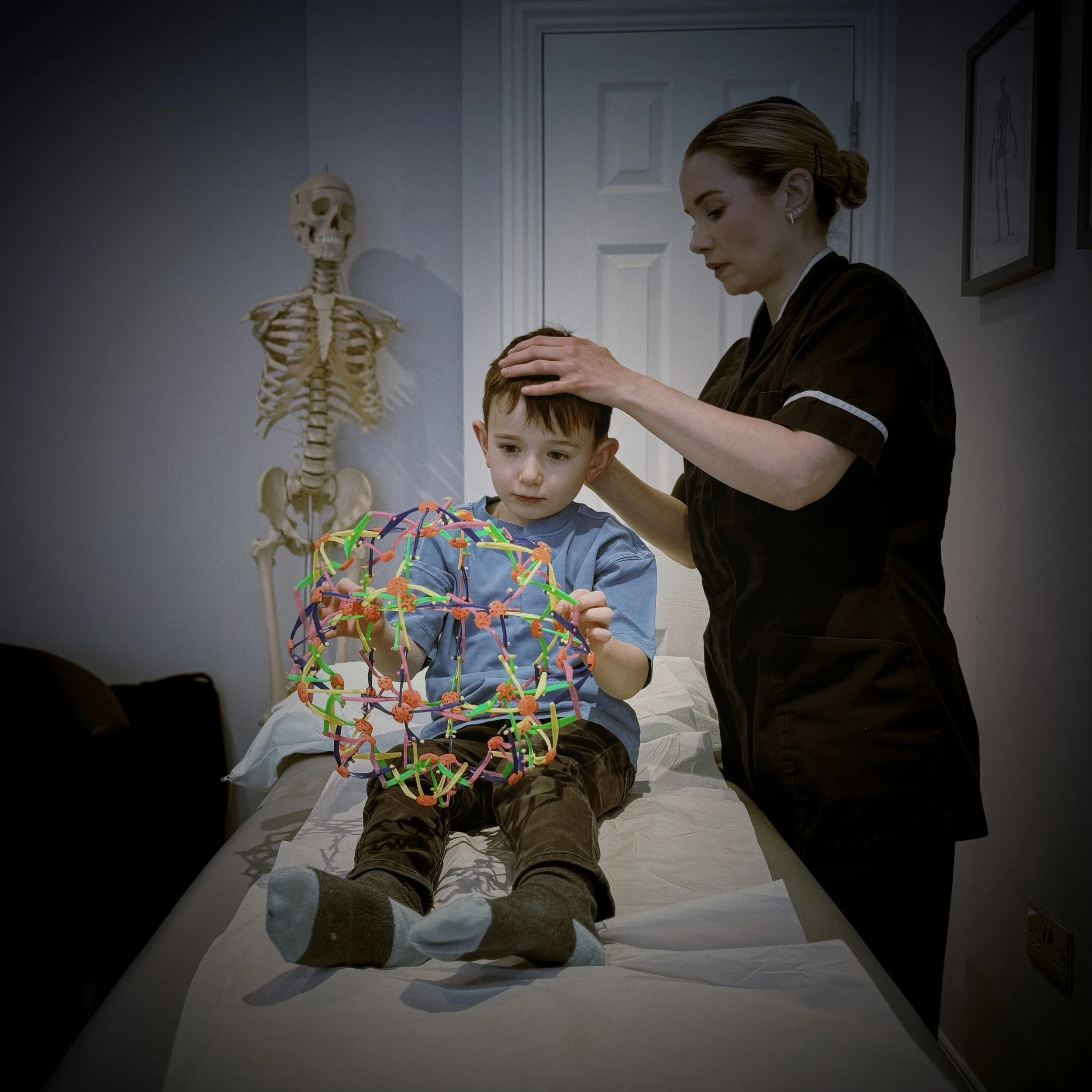 A woman in scrubs performs an examination or treatment on a young boy lying on a hospital bed, with a skeletal model in the background and framed artwork on the wall.