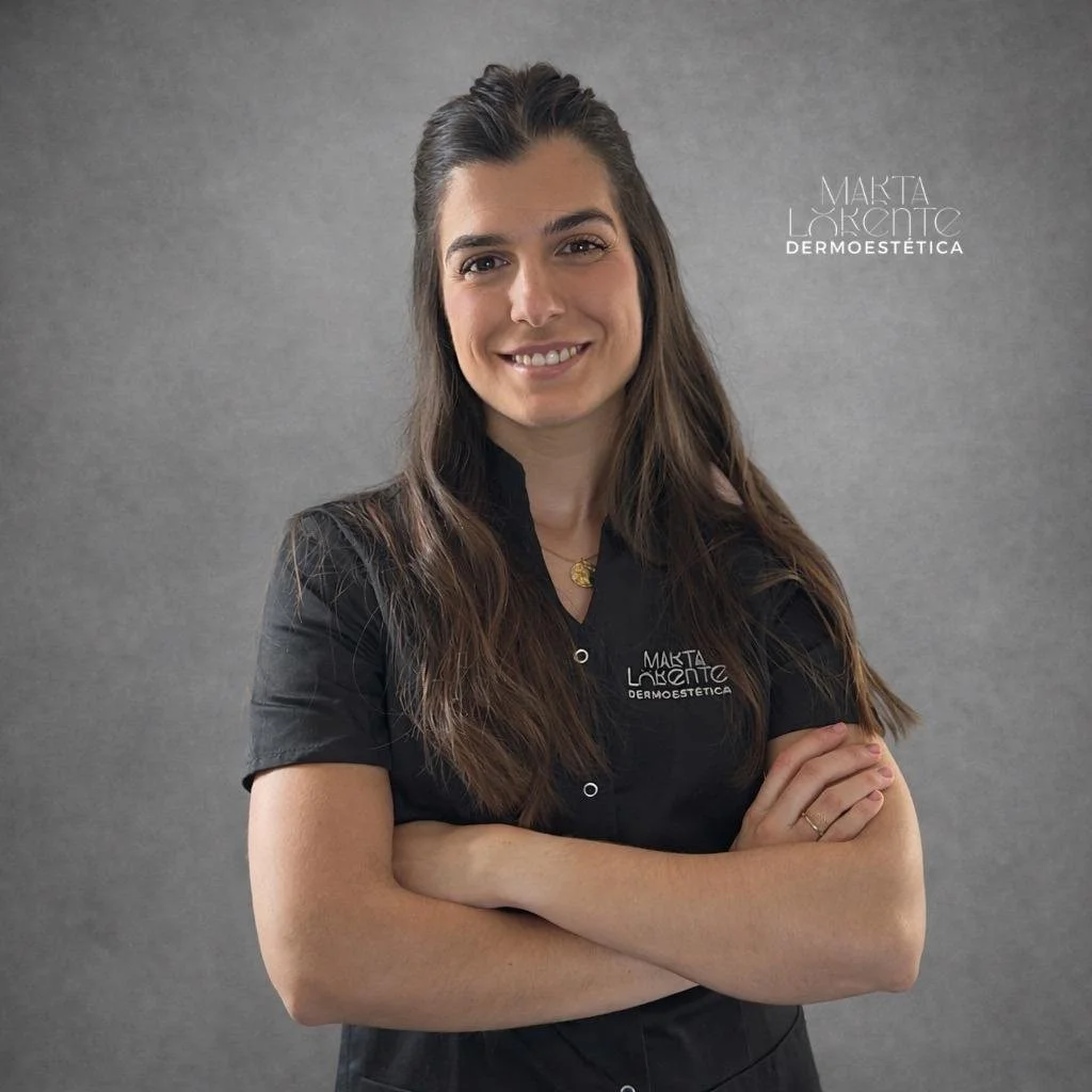 Mujer con cabello largo y oscuro, sonriendo y con brazos cruzados, vestida con uniforme negro de estética, sobre fondo gris