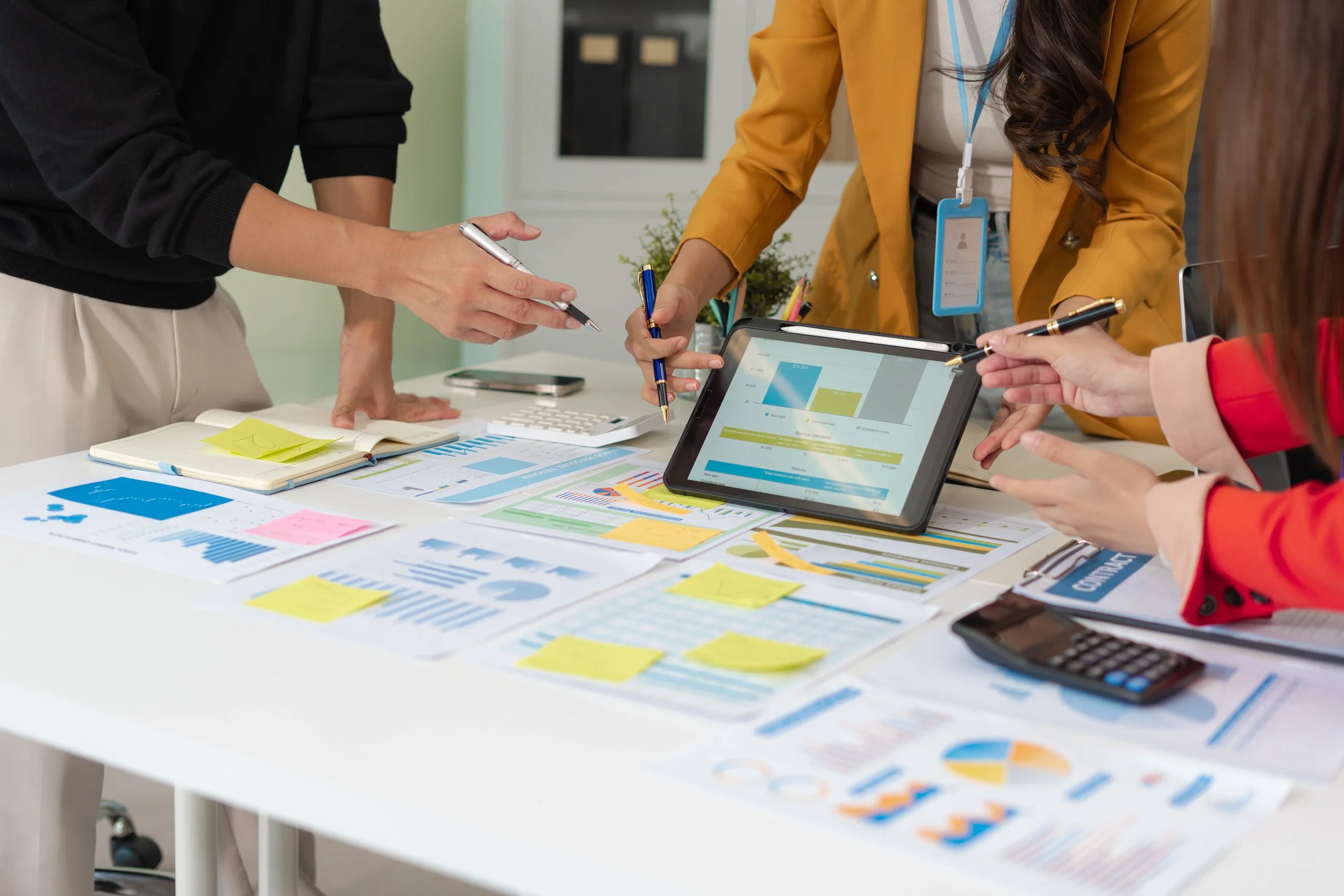 Group of people working together on a business project with charts, graphs, sticky notes, a tablet, and a calculator on the table.