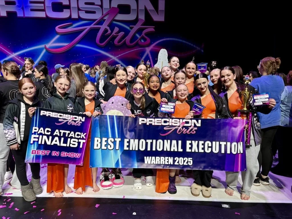 A group of young dancers holding awards and banners on stage after a dance competition. The background has a logo for Precision Arts, and the signs indicate they are finalists in Pac Attack and received the Best Emotional Execution award at Warren 20