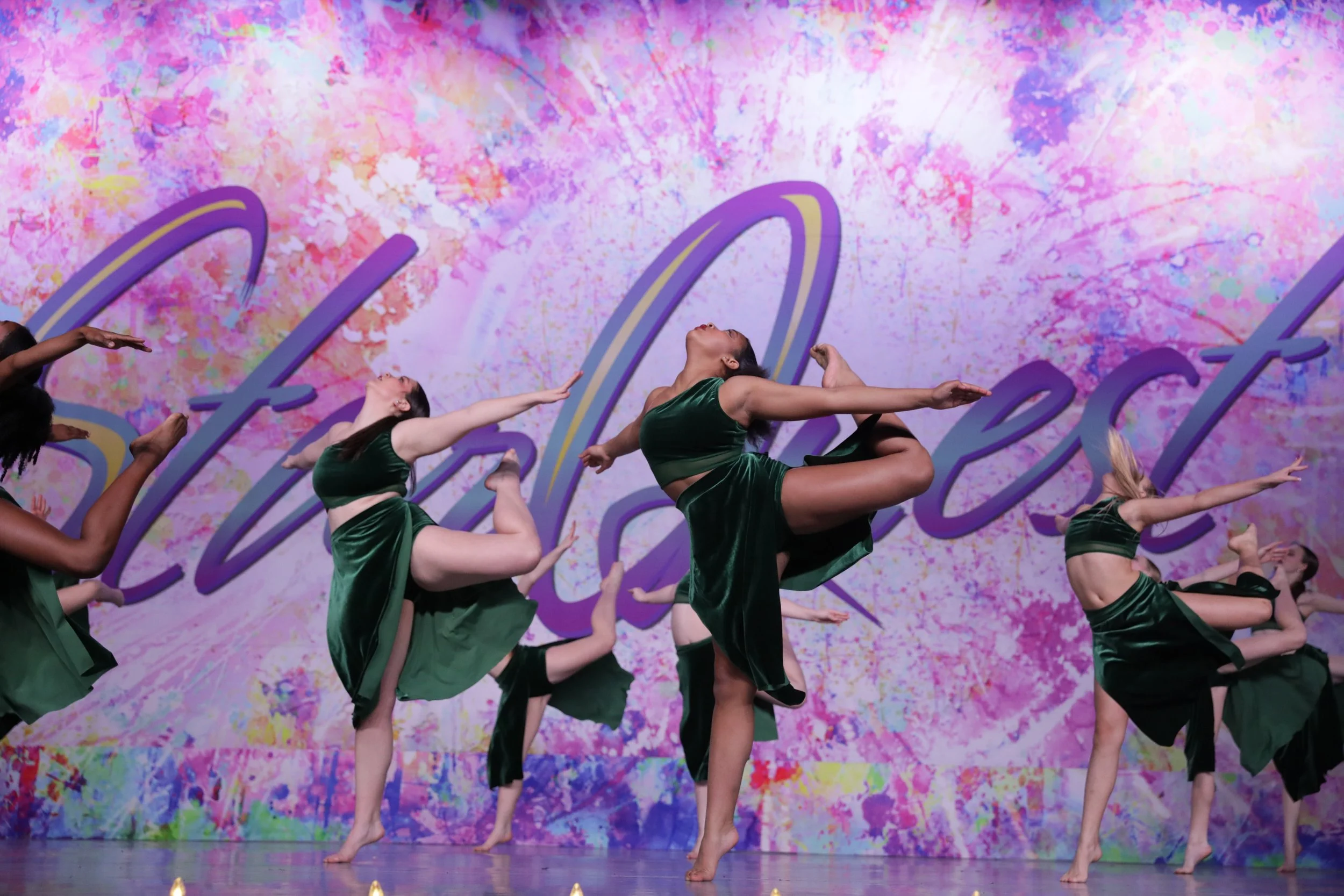 A group of dancers performing on stage with a colorful, Artistic backdrop. The dancers are wearing green costumes and are in various dance poses.