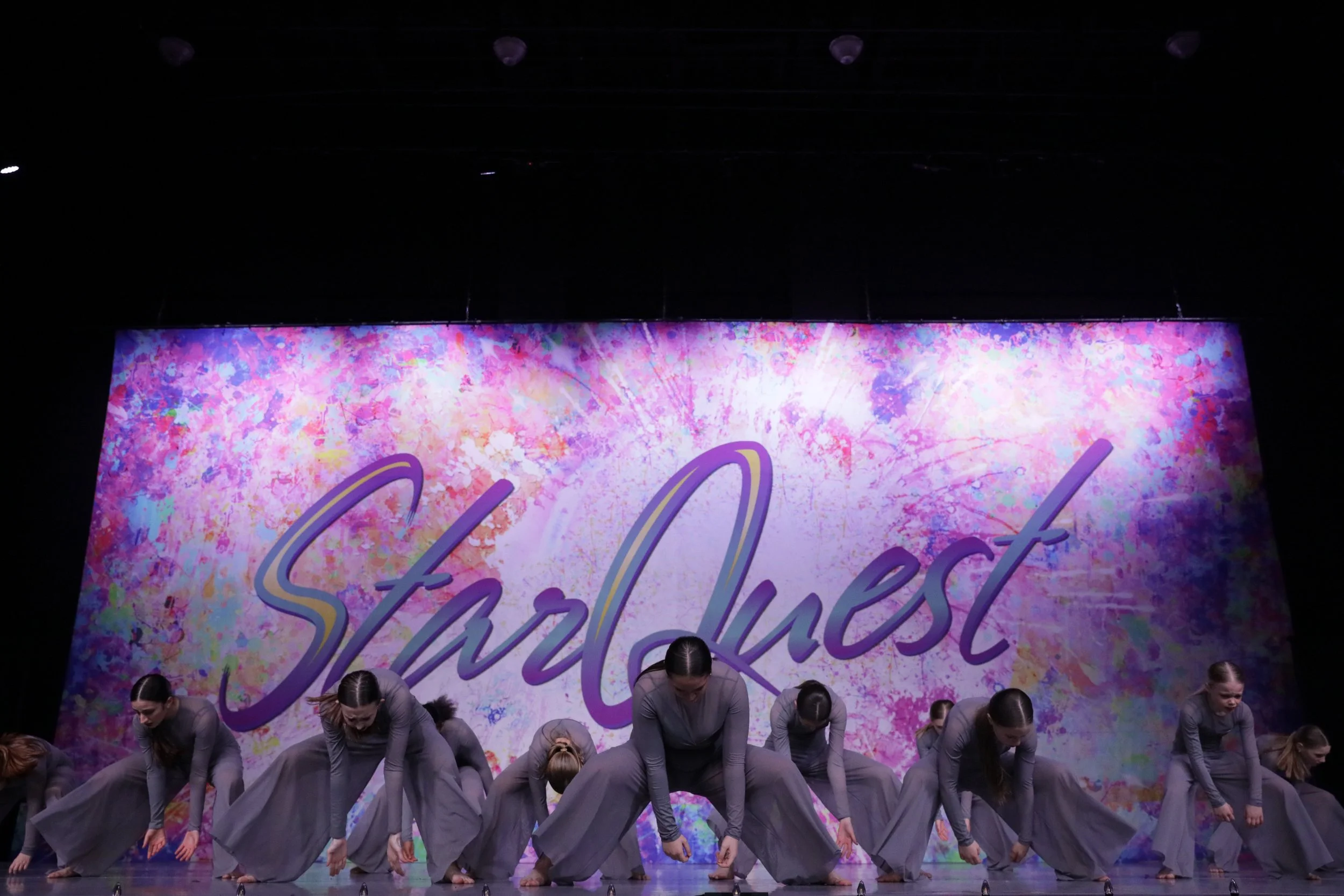 Group of dancers dressed in gray performing on stage in front of a colorful backdrop with the words "Starlust" written in purple.