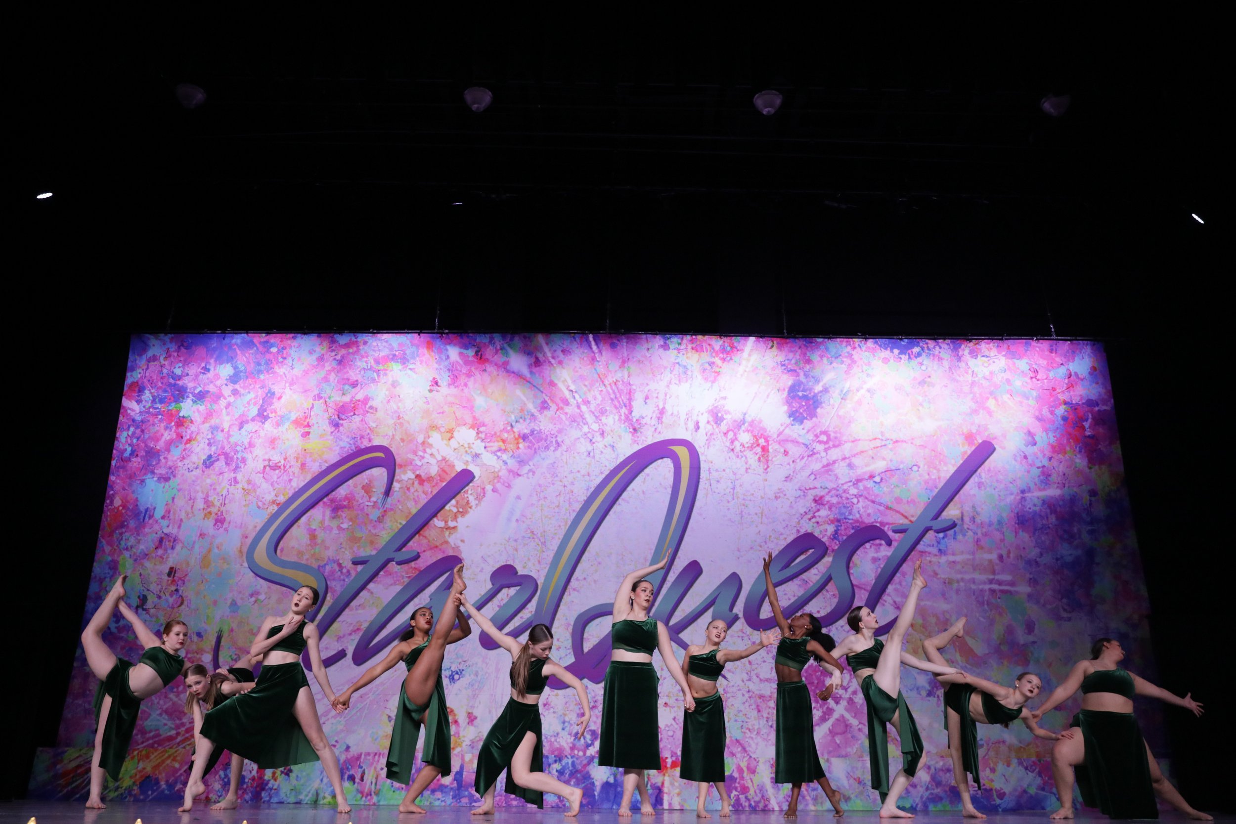 Dance performance on stage with ten dancers wearing dark green costumes, performing a pose in front of a colorful backdrop with the words 'StarQuest'.